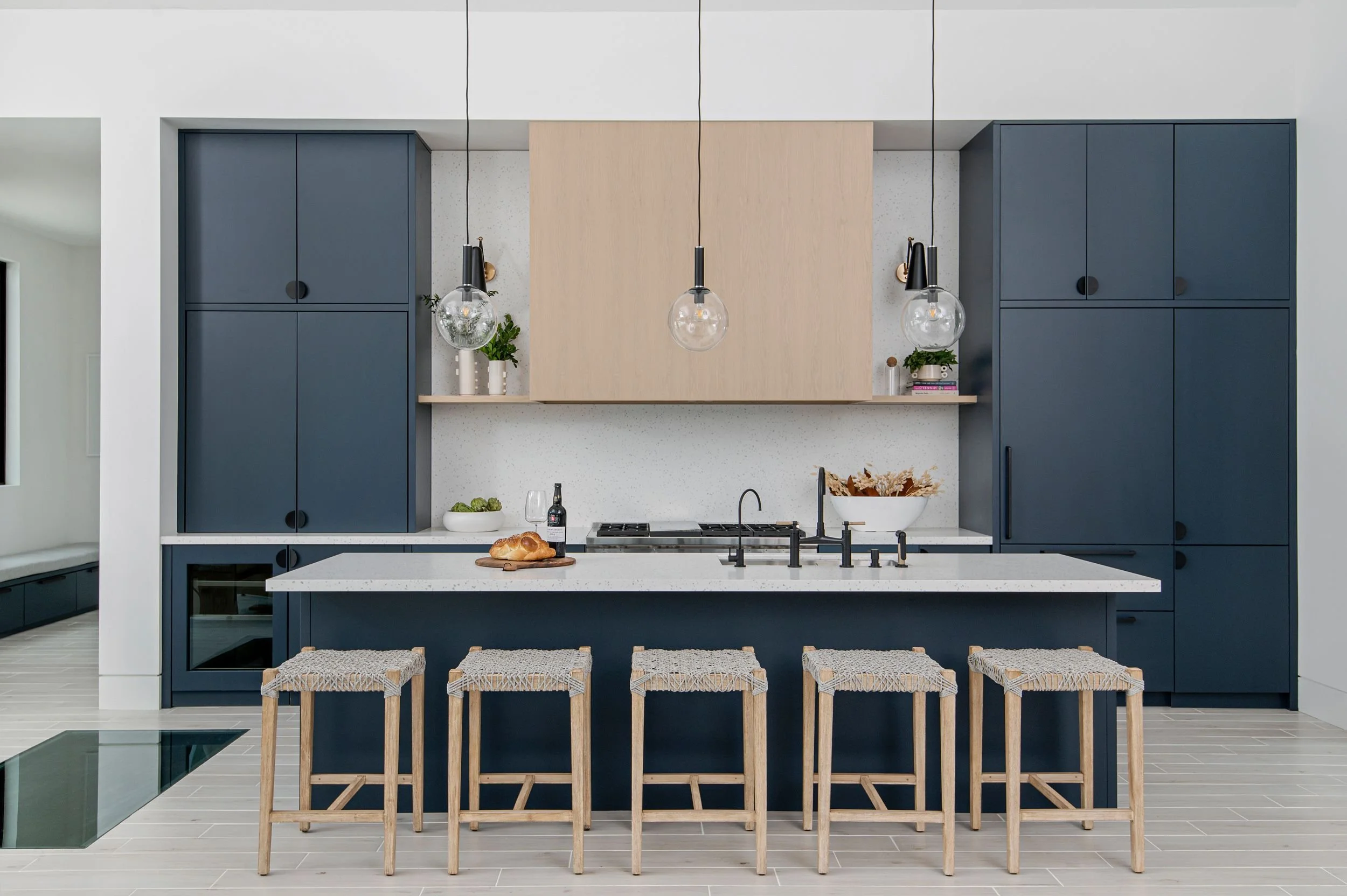 Modern kitchen with navy blue cabinets, white countertop island with five matching stools, pendant lights, and minimal decor.
