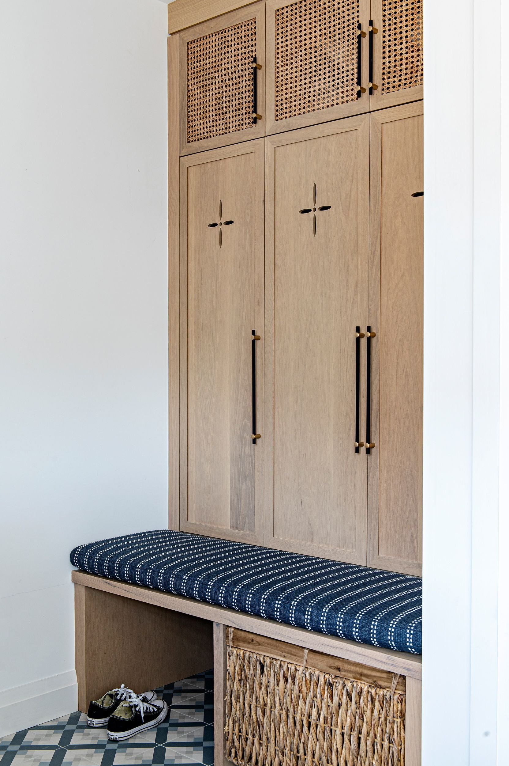 Built-in wooden cabinet with woven rattan upper doors, black and gold handles, and a padded bench with blue and white striped cushion below. There is a wicker storage basket underneath and black sneakers on a patterned tile floor.