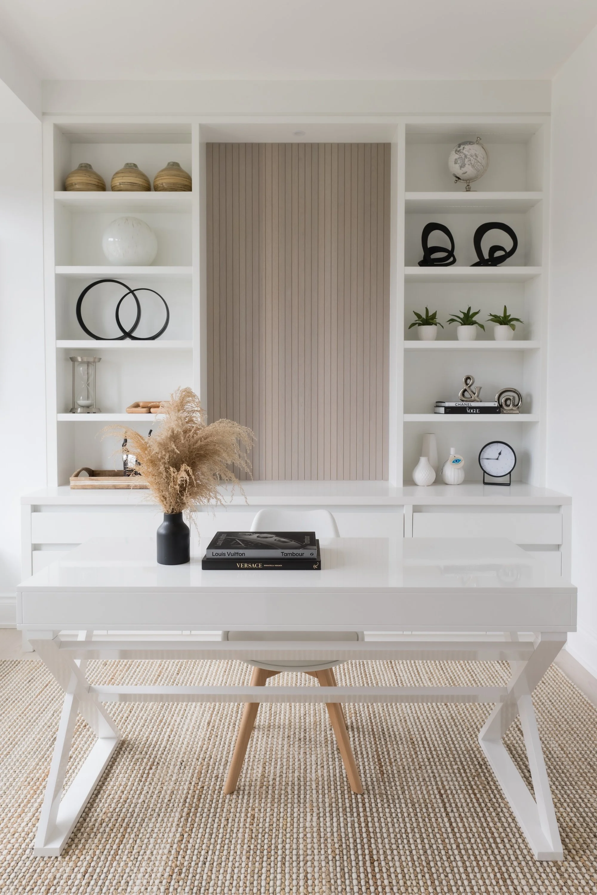 A modern, minimalist home office with white shelves. The shelves hold decorative objects like vases, books, and globes. A white desk with a black vase containing dried pampas grass, and a book on top. The room features neutral tones and a beige rug.