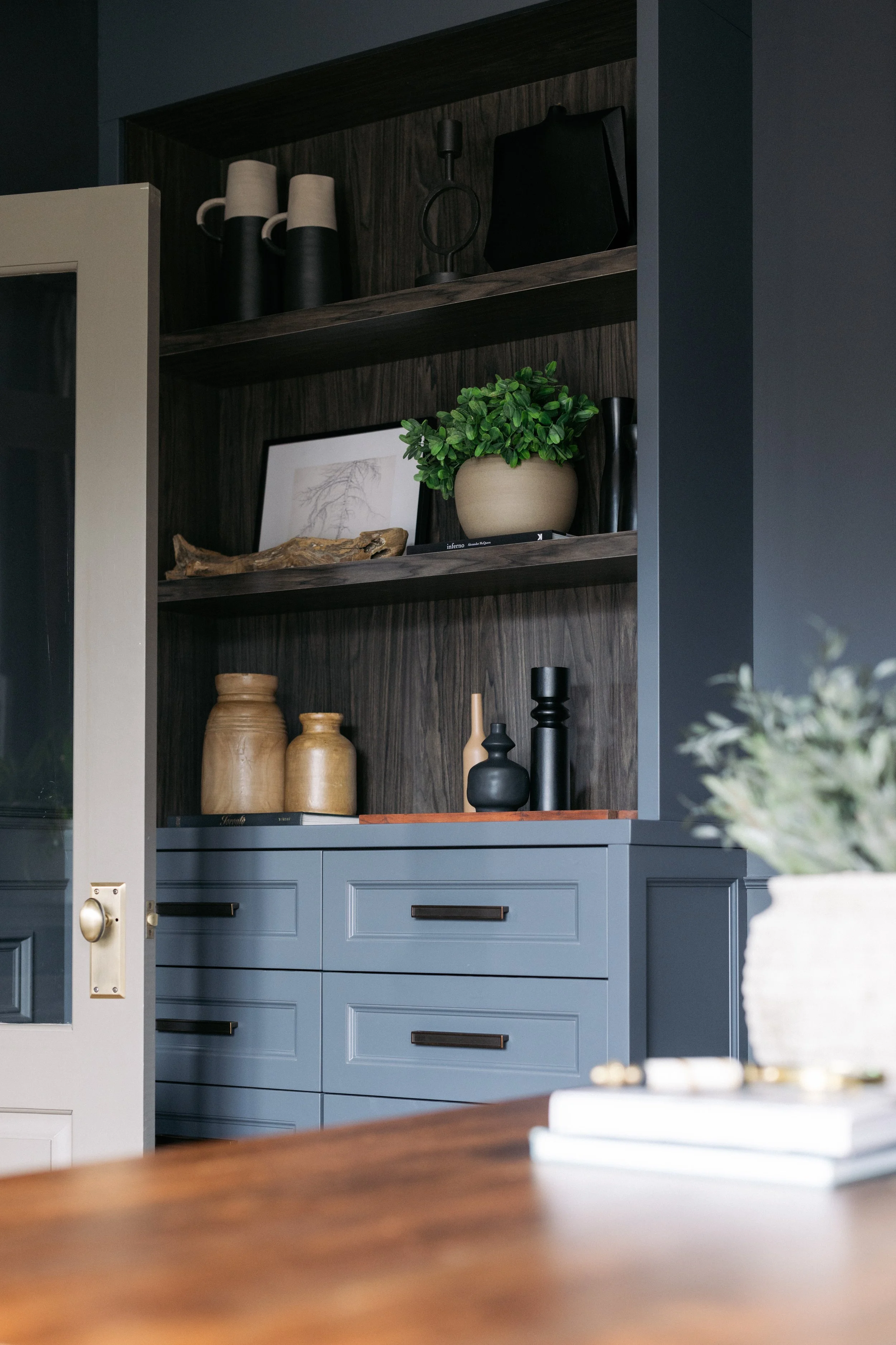 Wooden cabinetry with decorative vases, a plant in a beige pot, framed artwork, and various objects on open shelves.