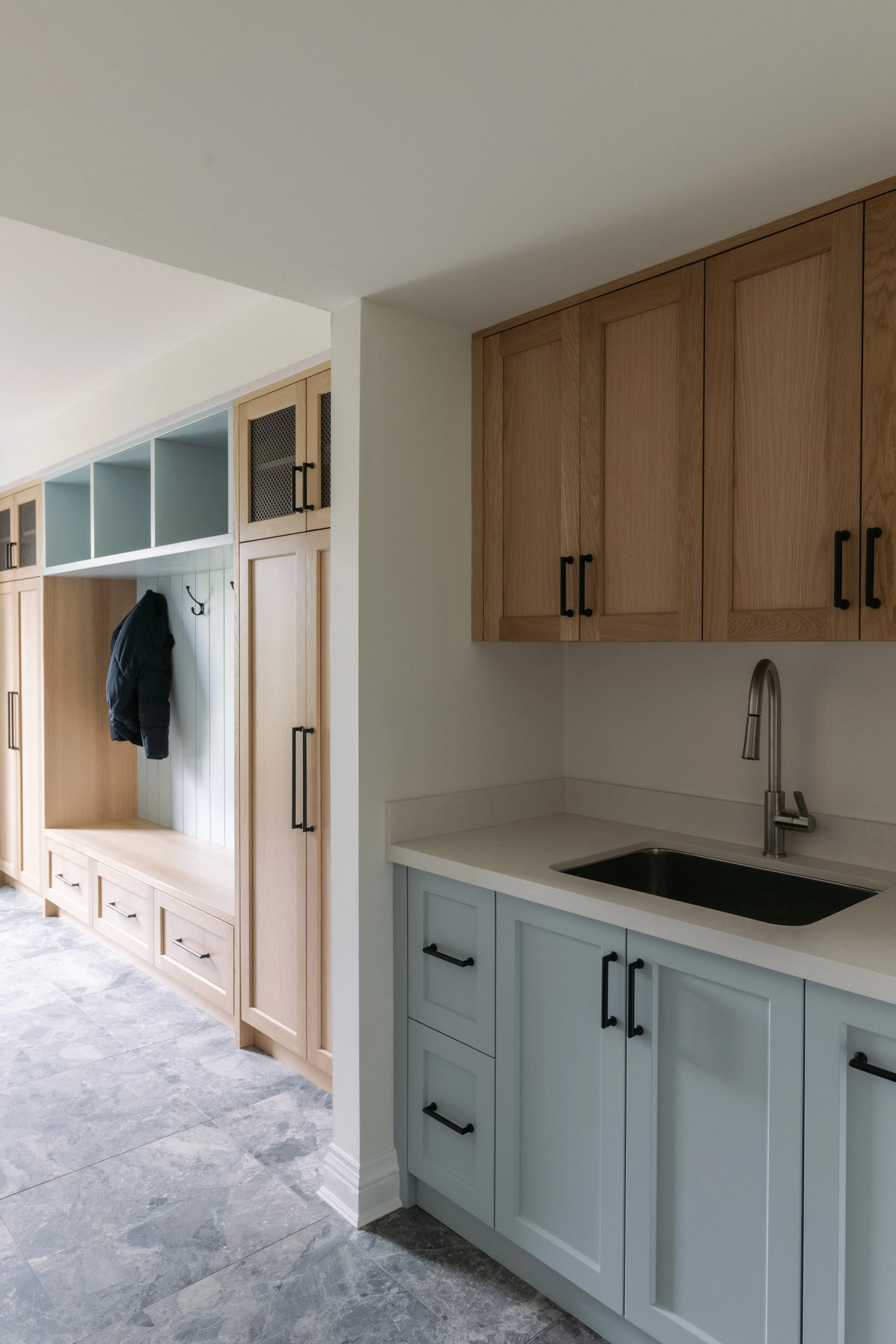 Interior view of a laundry room or mudroom with wooden cabinets, a small sink, and a hallway with built-in storage, coat hooks, and a bench.