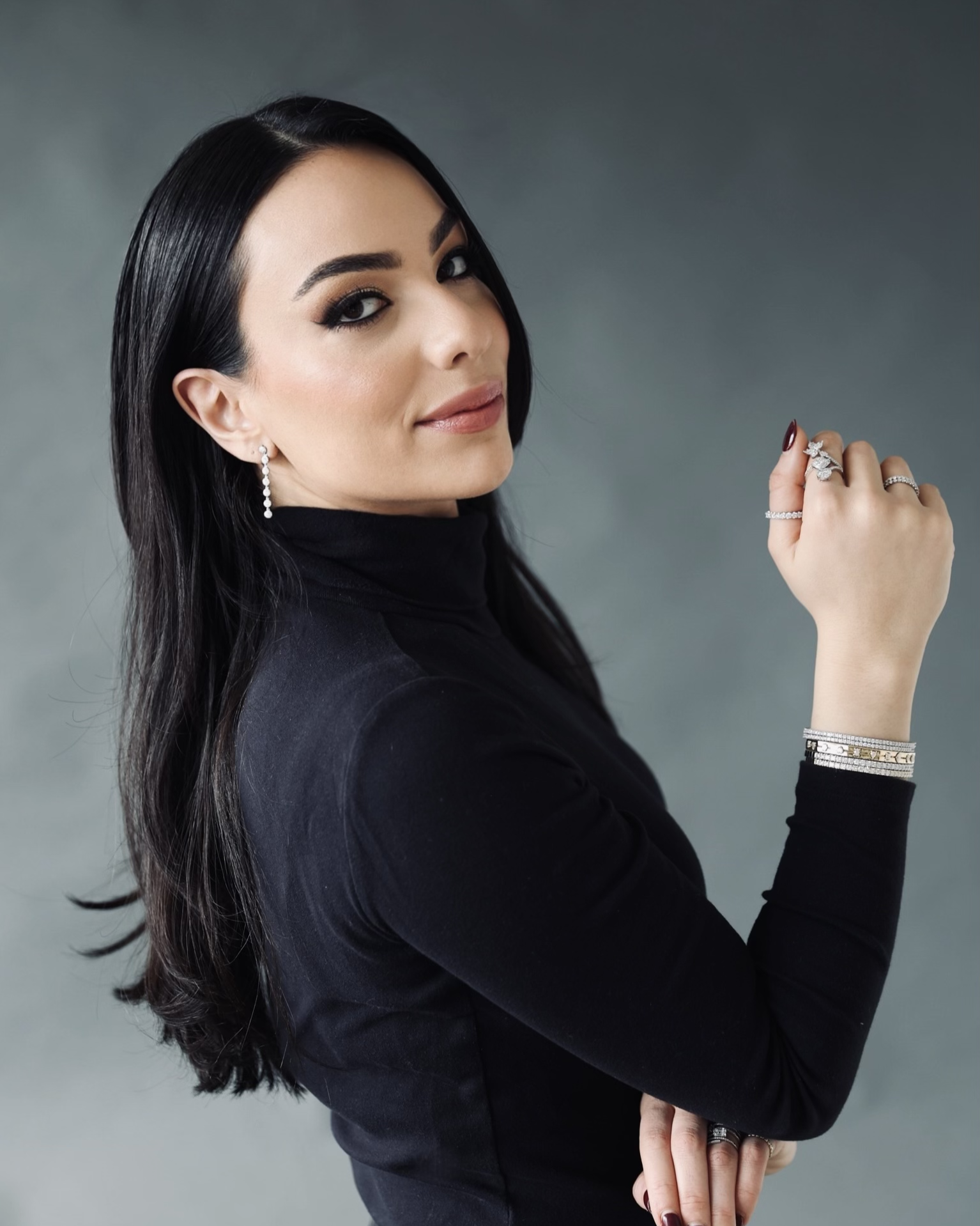 A woman with long dark hair, wearing a black turtleneck, earrings, and multiple rings and bracelets, looks at the camera with a confident expression against a grey background.