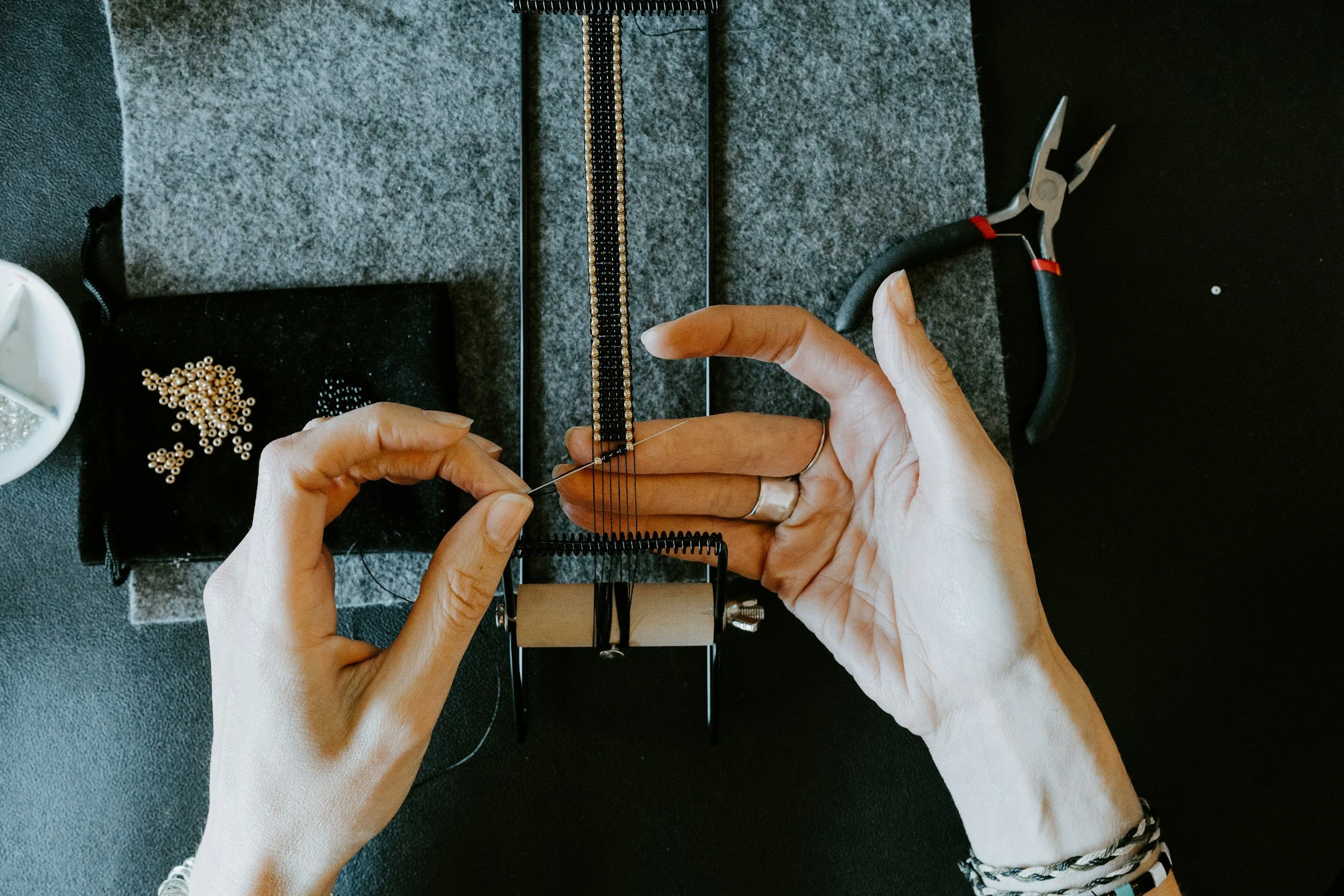 Person crafting jewelry with beads, using pliers on a black desk with tools and beads.