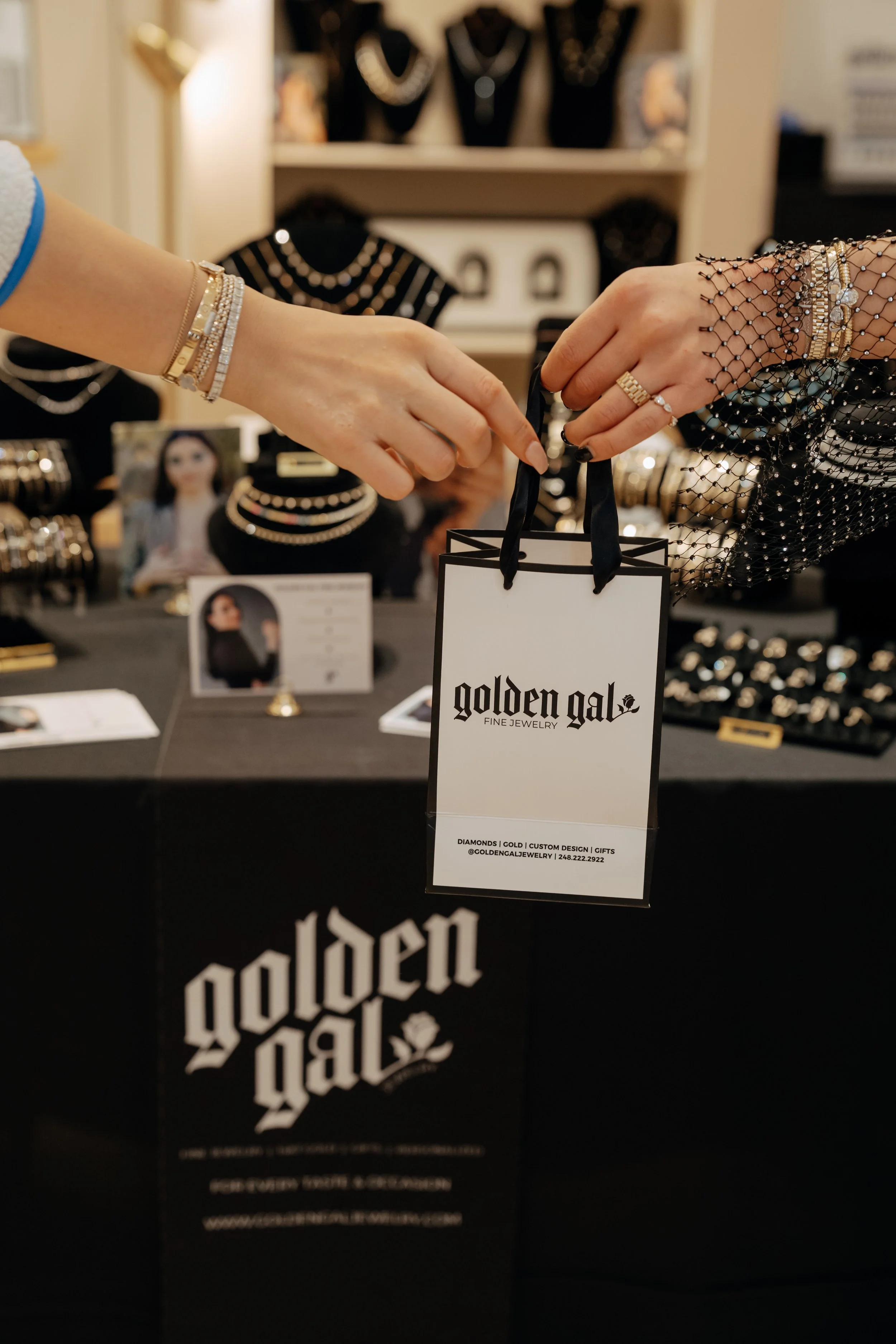 Two women exchanging a shopping gift bag from Golden Gal Fine Jewelry at a jewelry booth, with jewelry displays and photos in the background.