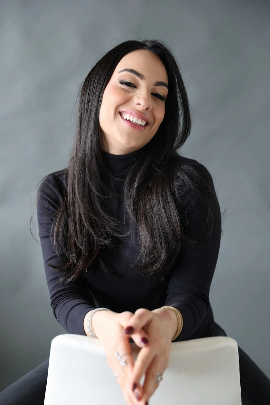 A woman with long dark hair, smiling, wearing a black turtleneck, sitting with her hands clasped, in front of a plain gray background.