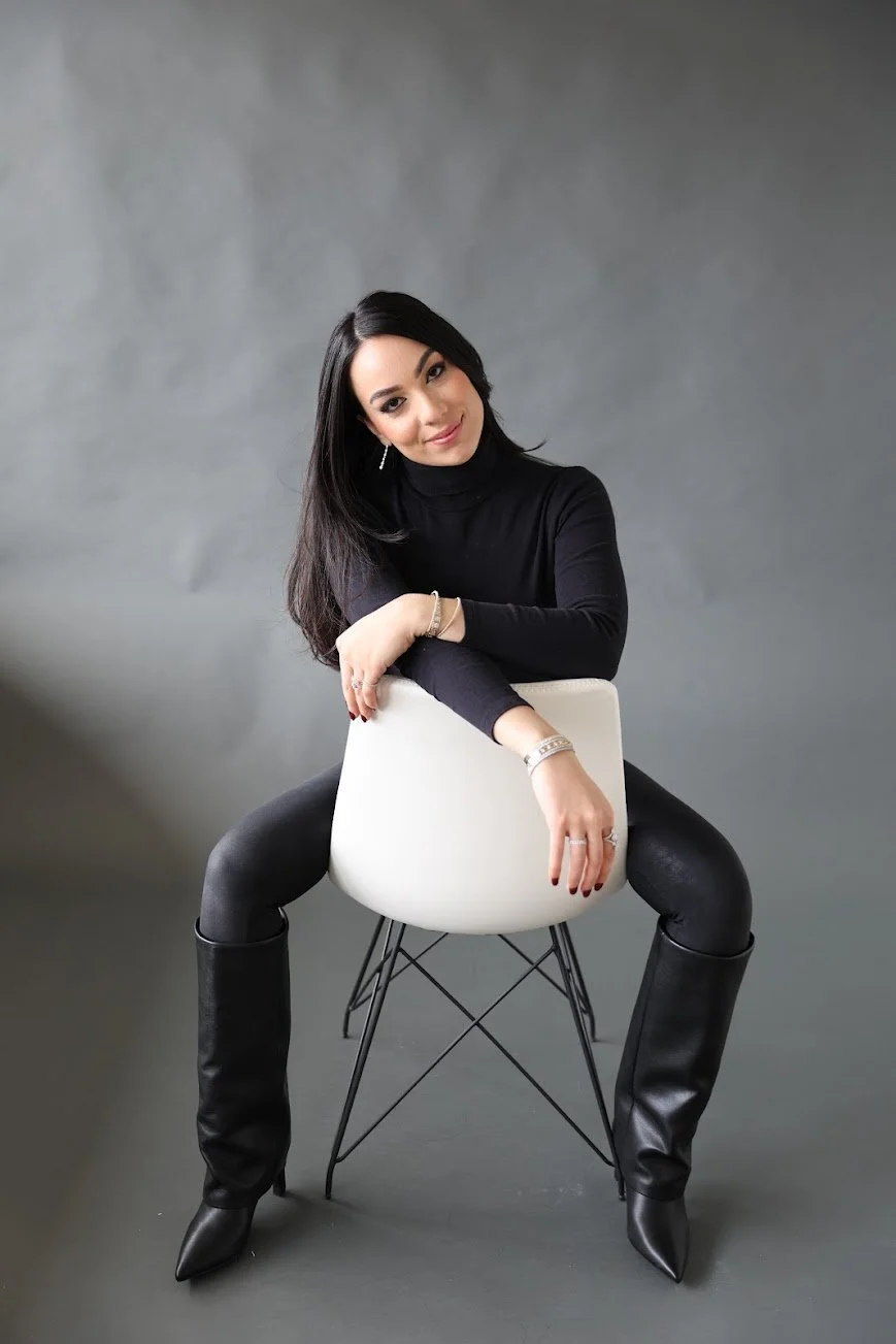 A woman with long dark hair wearing a black turtleneck and shiny black pants, sitting on a white chair against a gray background.