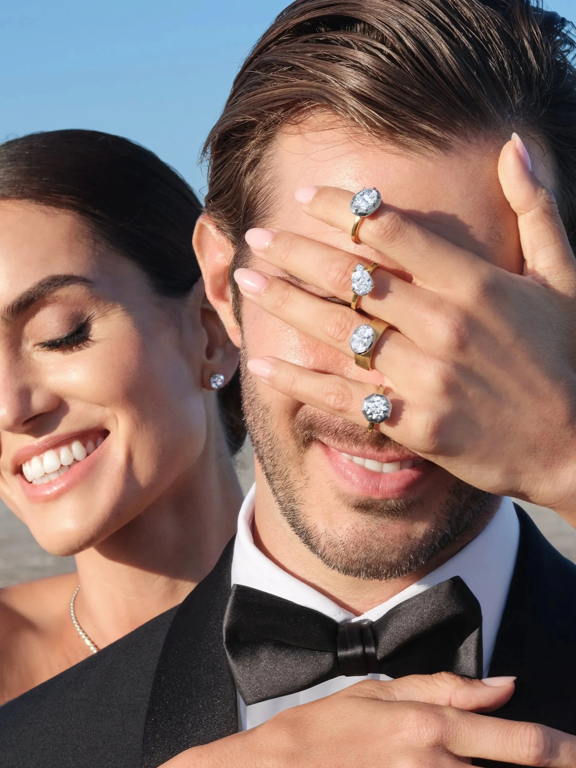 A smiling woman in formal attire, wearing diamond earrings and an elegant necklace, standing next to a man wearing a tuxedo and bow tie. The woman is covering one of the man's eyes with her hand, showcasing multiple rings with large gemstones on her fingers.