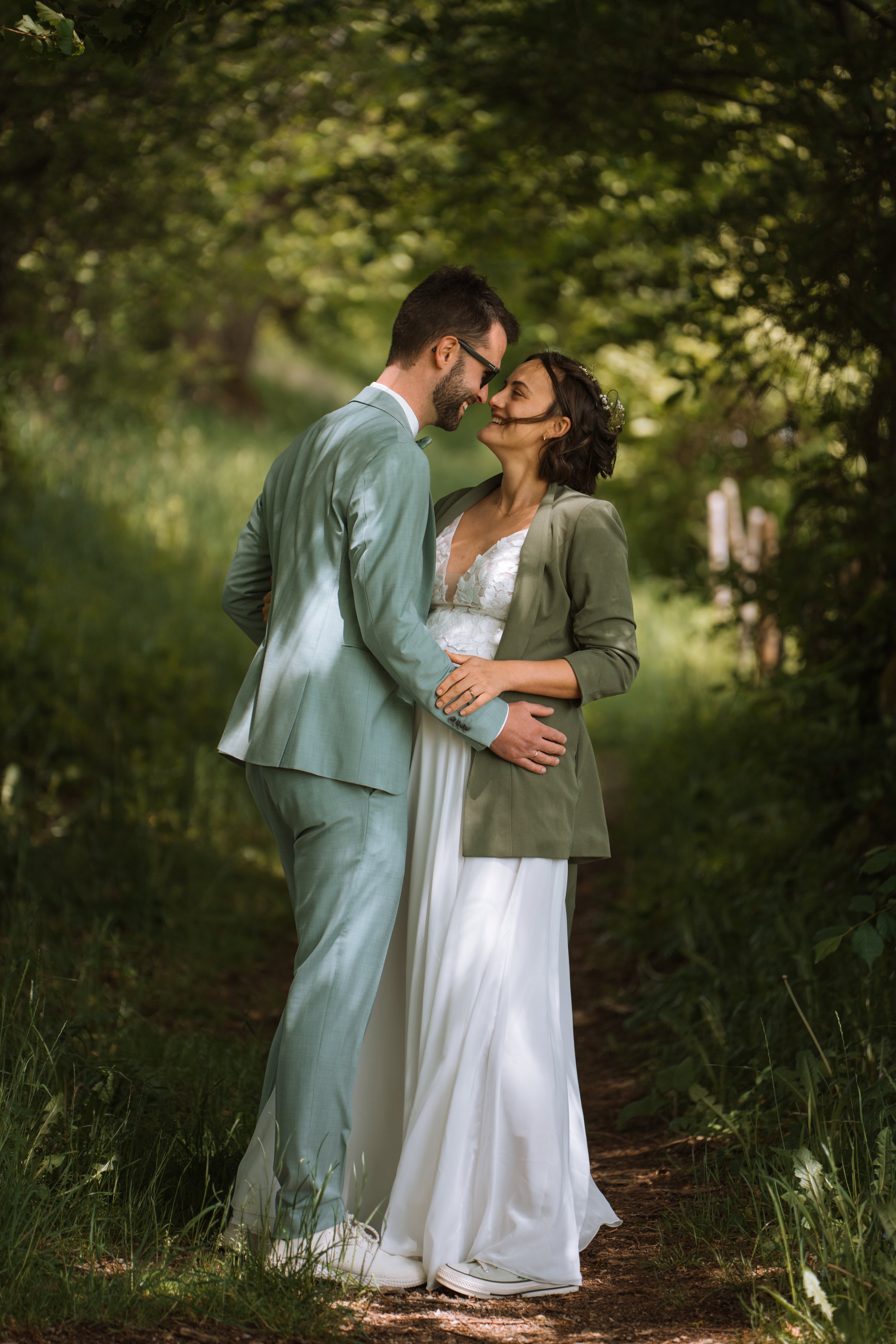 Ein Paar in Hochzeitskleidung steht Rücken an Rücken auf einem Waldblick, lächelt sich an und berührt sich zärtlich.