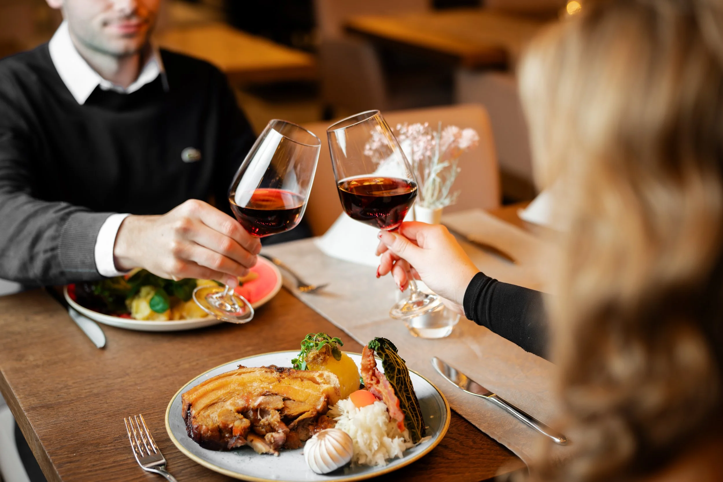 Zwei Personen stoßen sich mit Rotweingläsern während eines Abendessens im Restaurant zu. Auf dem Tisch stehen fertig angerichtete Mahlzeiten, darunter Schweinebraten mit Beilagen und ein Salat.