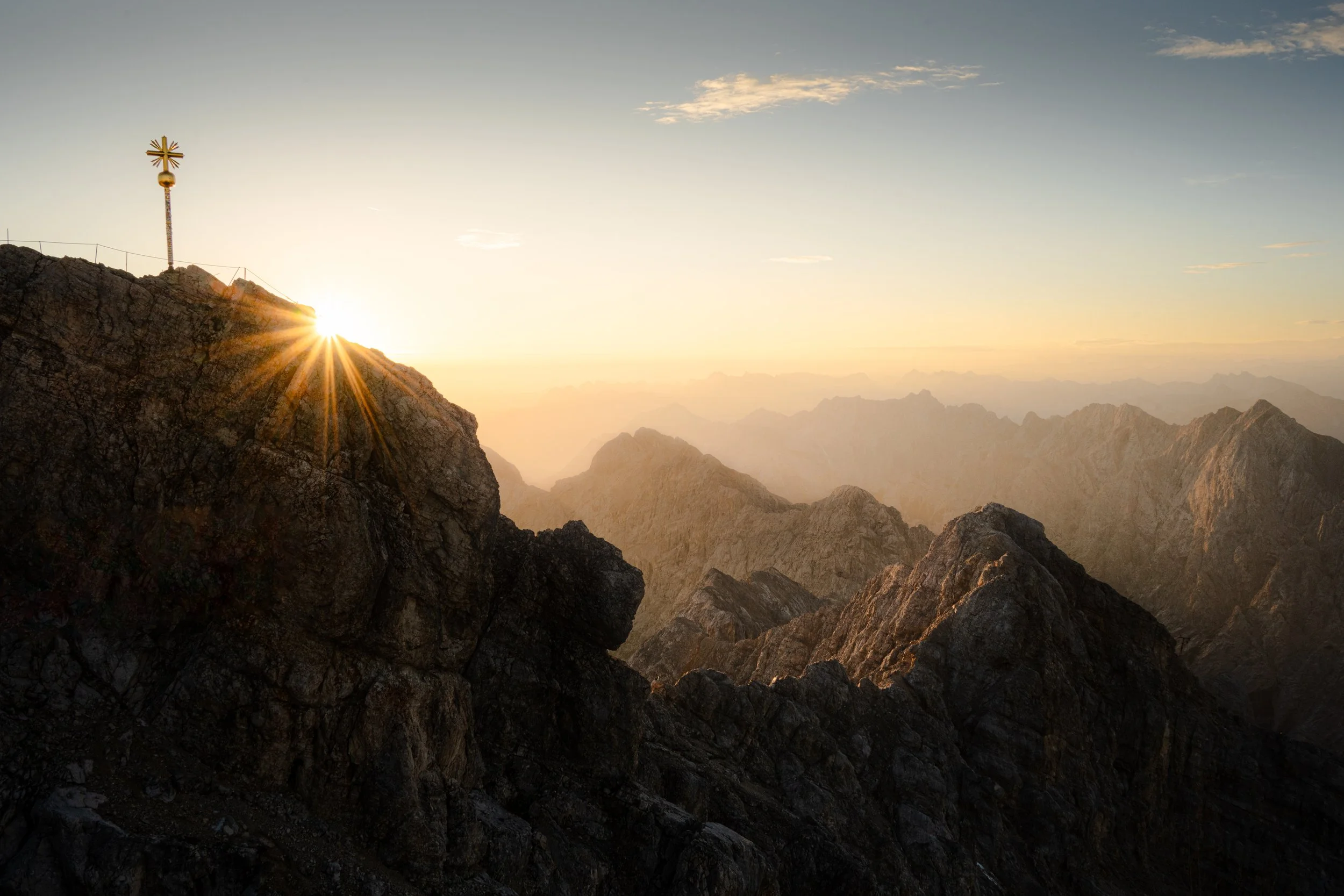 Berggipfel mit Kreuz und Sonnenschein, im Hintergrund Gebirge bei Sonnenaufgang oder Sonnenuntergang.