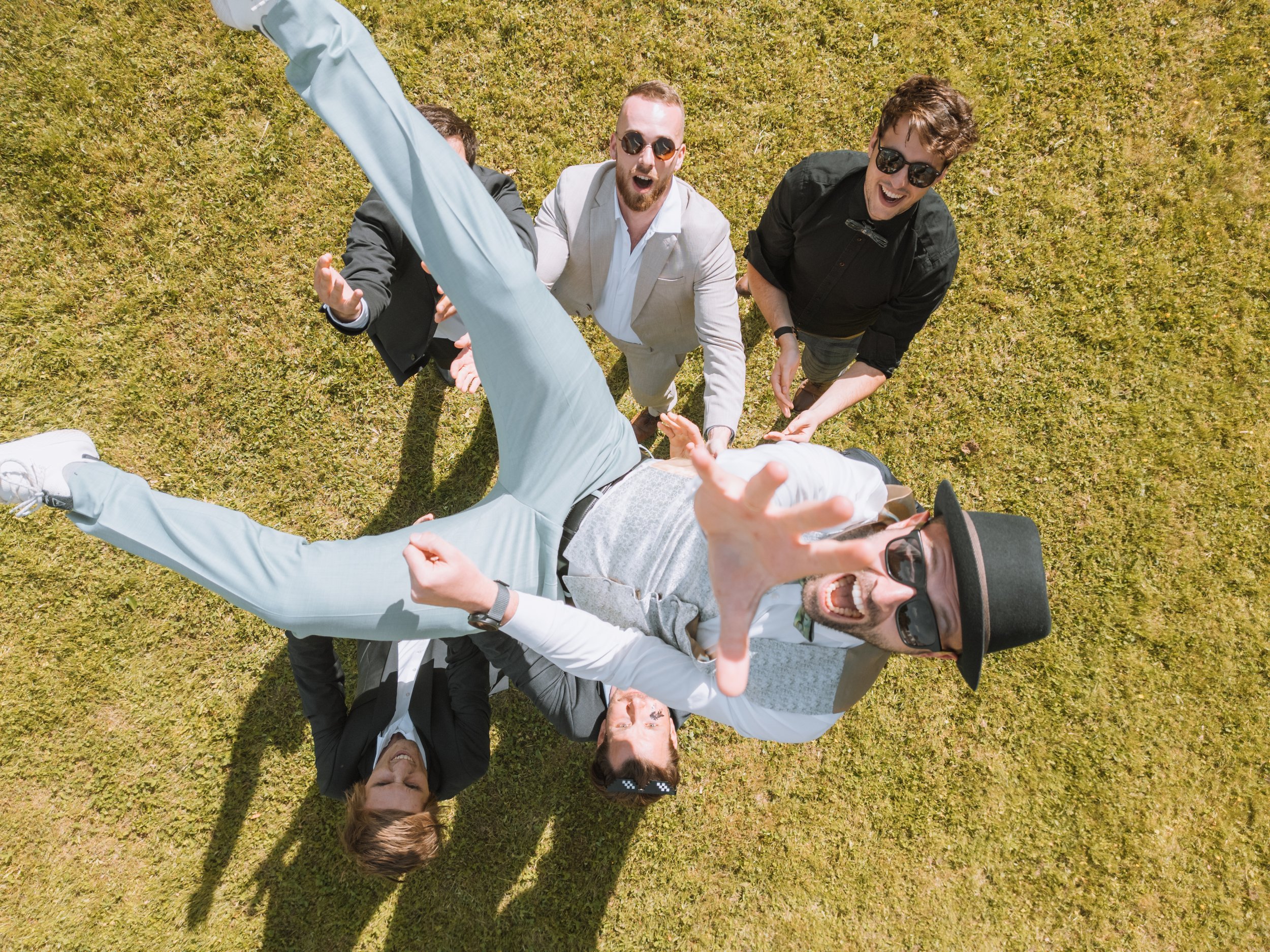 Eine Gruppe von fünf Menschen liegt auf dem Rasen, wobei vier von ihnen auf dem Boden sitzen oder liegen, und einer steht vorne mit einem Hut und Sonnenbrille. Sie lachen und scheinen Spaß zu haben.