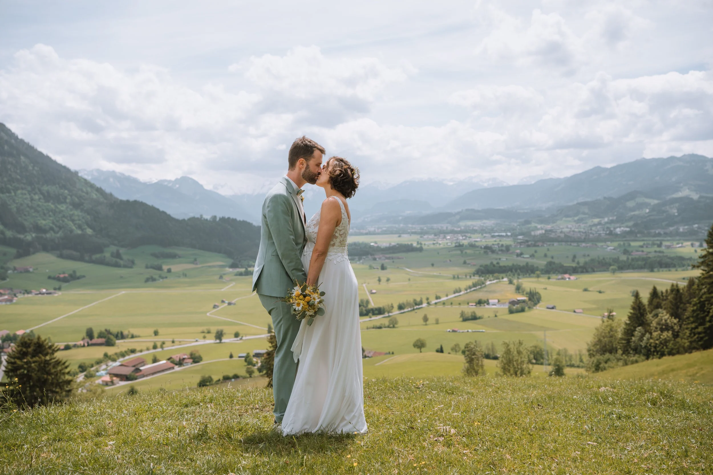 Ein Brautpaar küsst sich auf einer grünen Wiese vor einer malerischen Bergkulisse, mit Wolken am Himmel und einer weiten Landschaft im Hintergrund.