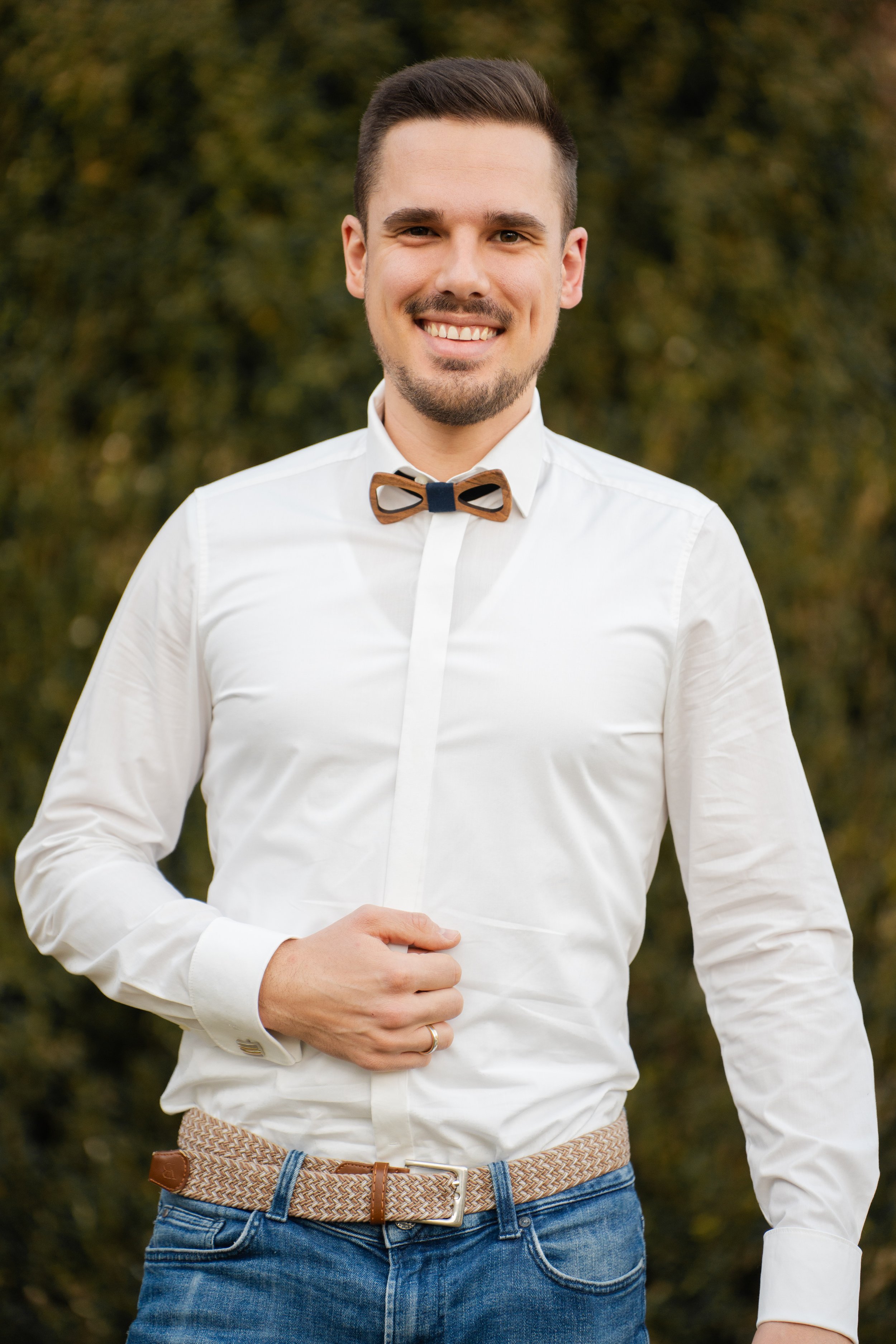 Ein Mann trägt eine weiße Hemd, eine braune Fliege und eine Denim-Hose. Er steht im Freien vor einem Baum mit Herbstlaub.