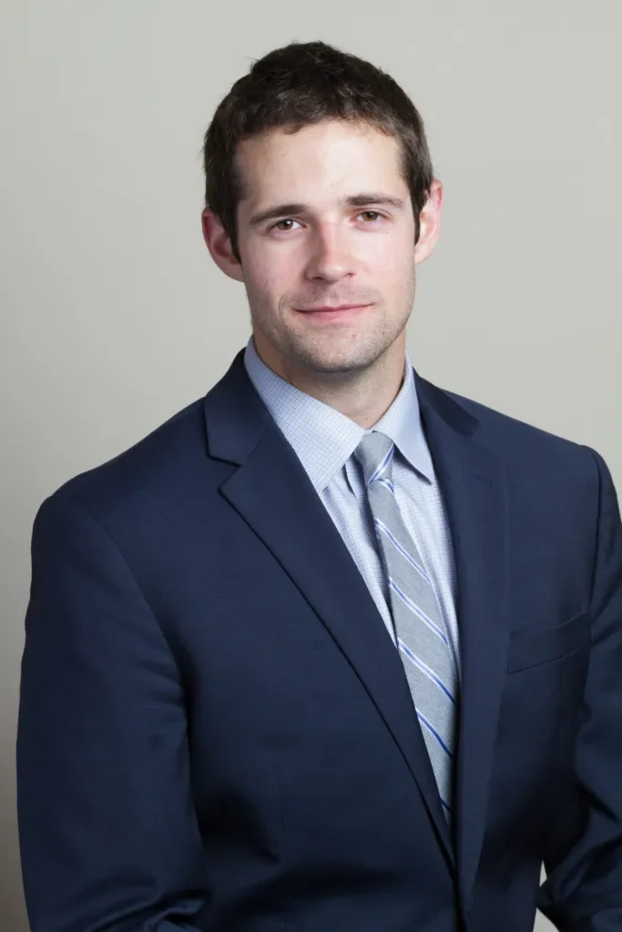Ryan Benson Tax Professional portrait of a young man in a dark blue suit, light blue dress shirt, and striped tie, standing against a plain light gray background.