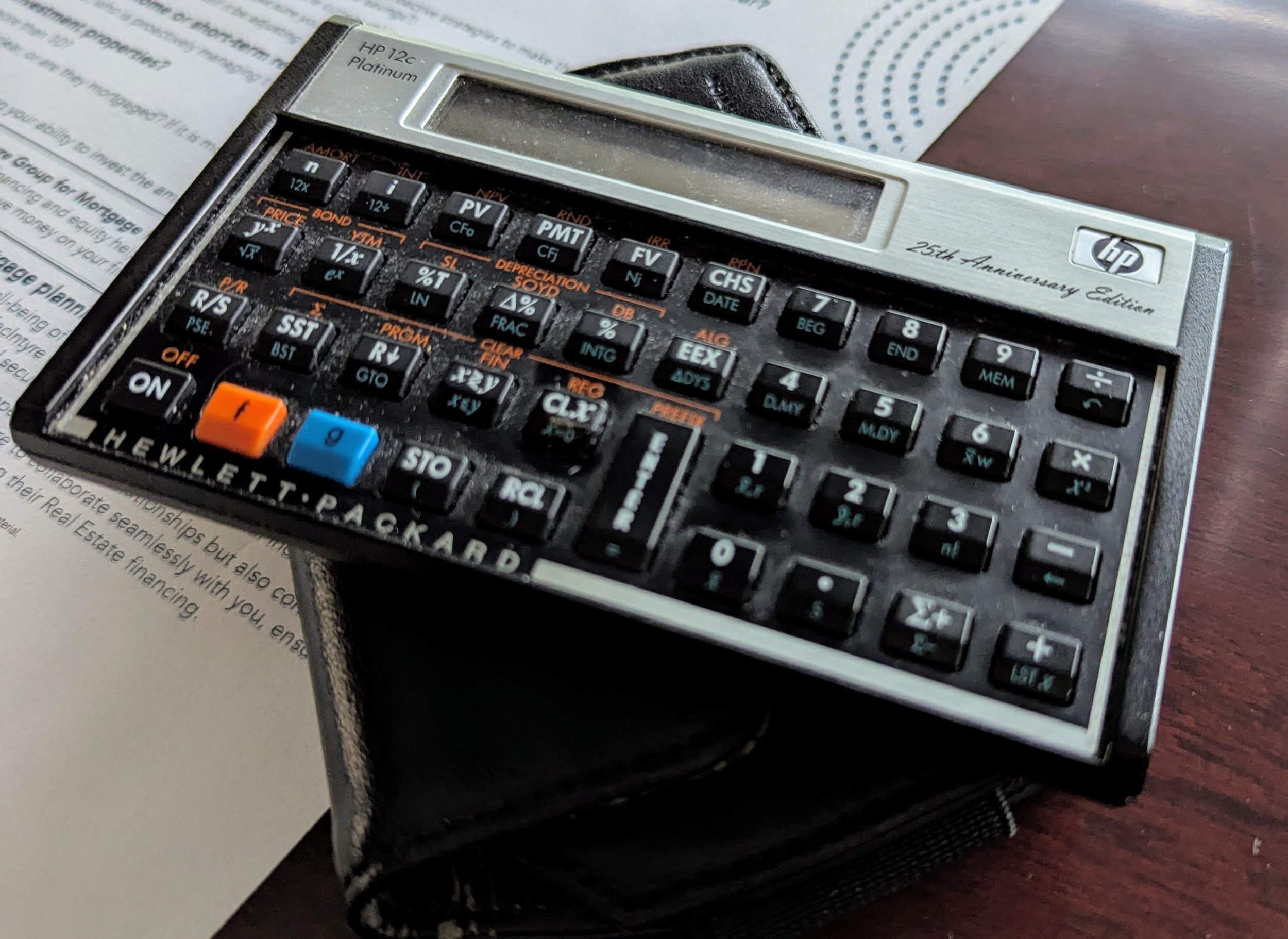 An HP 12c Platinum financial calculator resting on top of a black case, placed on a wooden surface. Partially visible paper is underneath the calculator.