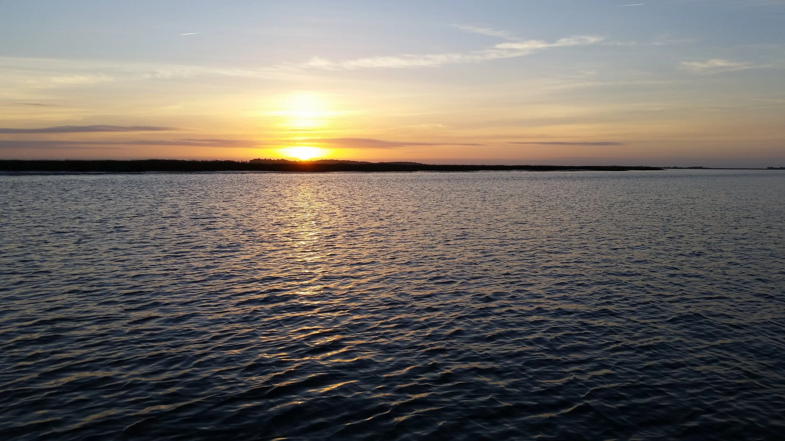 Sunrise over the Chesapeake Bay in Virginia with a distant shoreline and a partly cloudy sky.