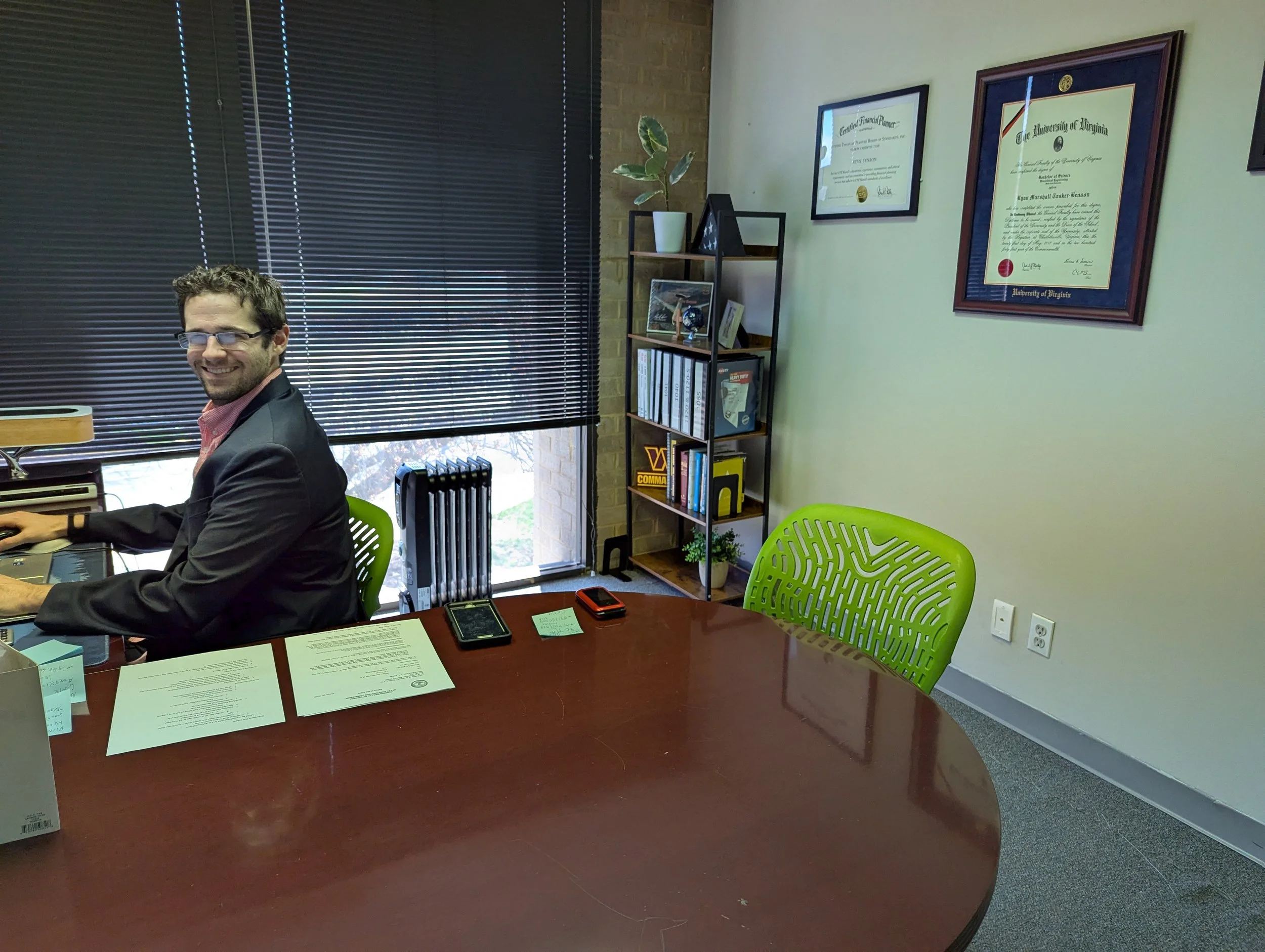 An office with a man sitting at a large wooden desk, smiling at the camera. There are documents, a smartphone, and office supplies on the desk. Taxes are being filed.