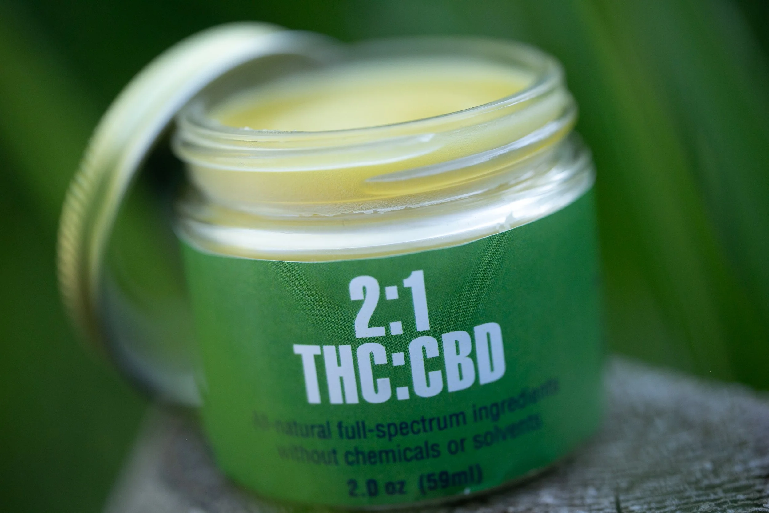 Glass jar labeled 2:1 THC:CBD ointment, opened on a wooden surface.