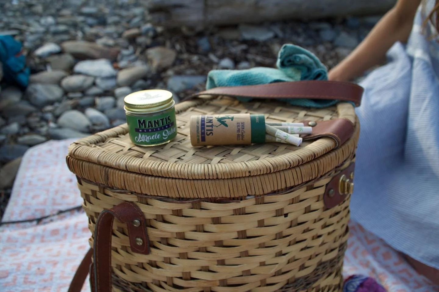 Wicker picnic basket with a jar of salve and tube on top, picnic blanket, and person sitting nearby, rocky ground background.