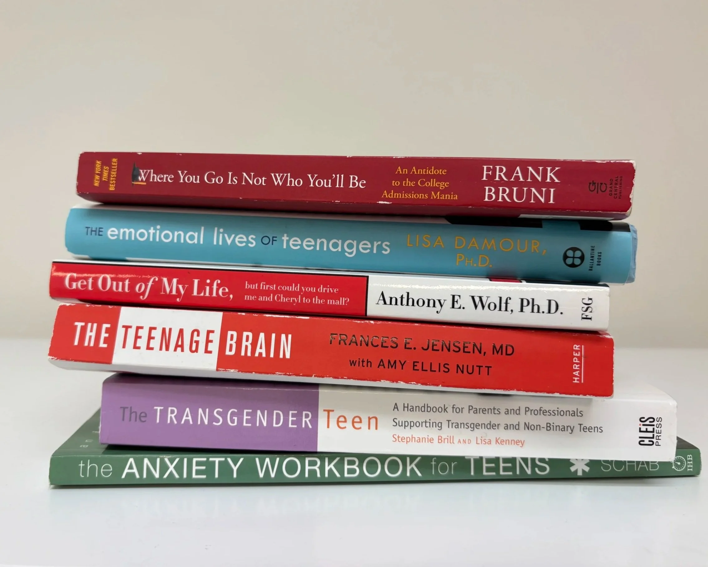Stack of seven books on a white surface against a light background. The titles from top to bottom are: 'Where You Go Is Not Who You’ll Be' by Frank Bruni, 'The Emotional Lives of Teenagers' by Lisa Damour, 'Get Out of My Life' by Anthony E. Wolf, 'The Teenage Brain' by Frances E. Jensen with Amy Ellis Nutt, 'The Transgender Teen' by Stephanie Brill and Lisa Kenney, and 'The Anxiety Workbook for Teens' by Schab.