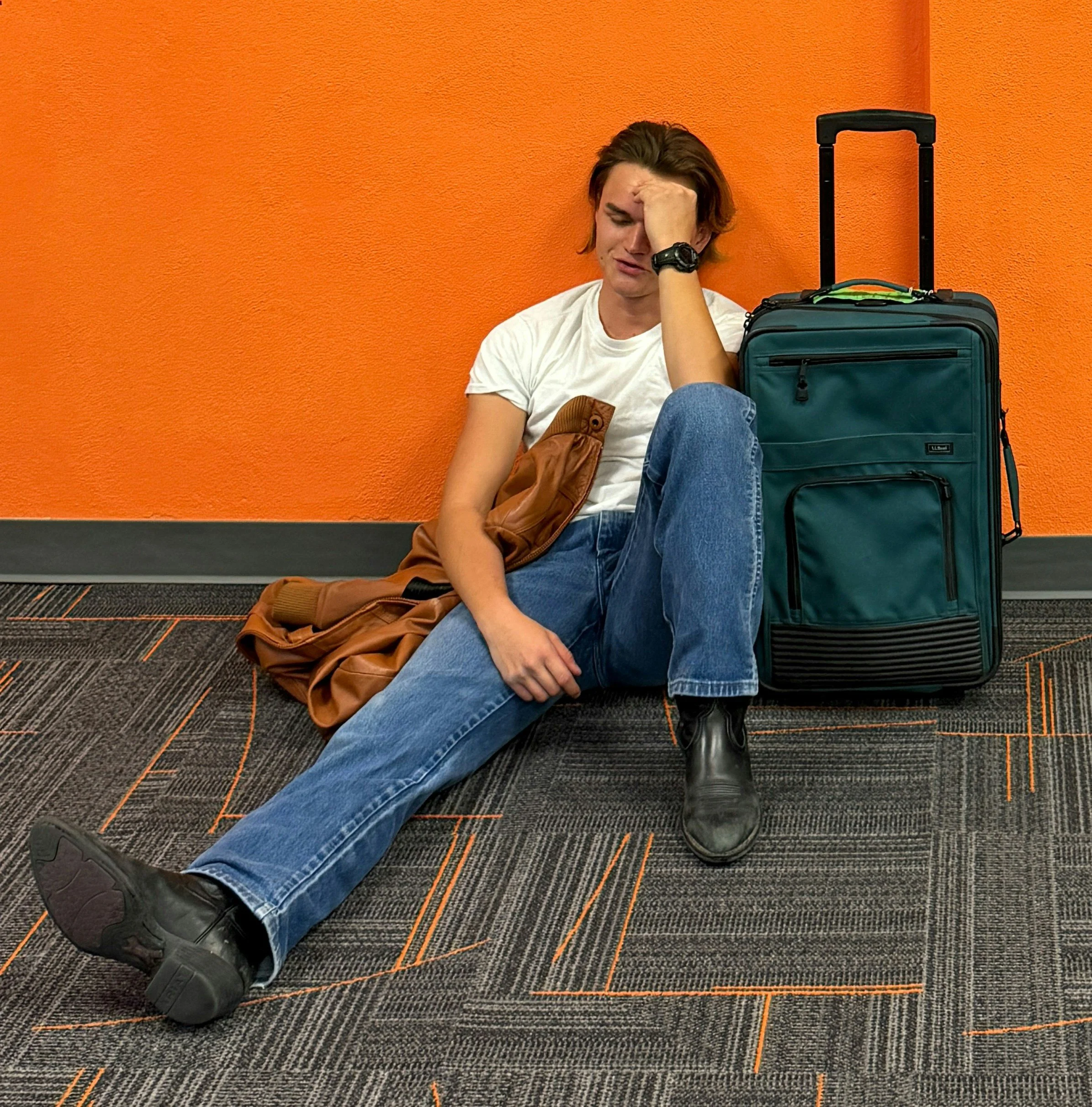 A young man sitting on the floor against an orange wall, appearing distressed, with a large teal suitcase and a brown jacket next to him.