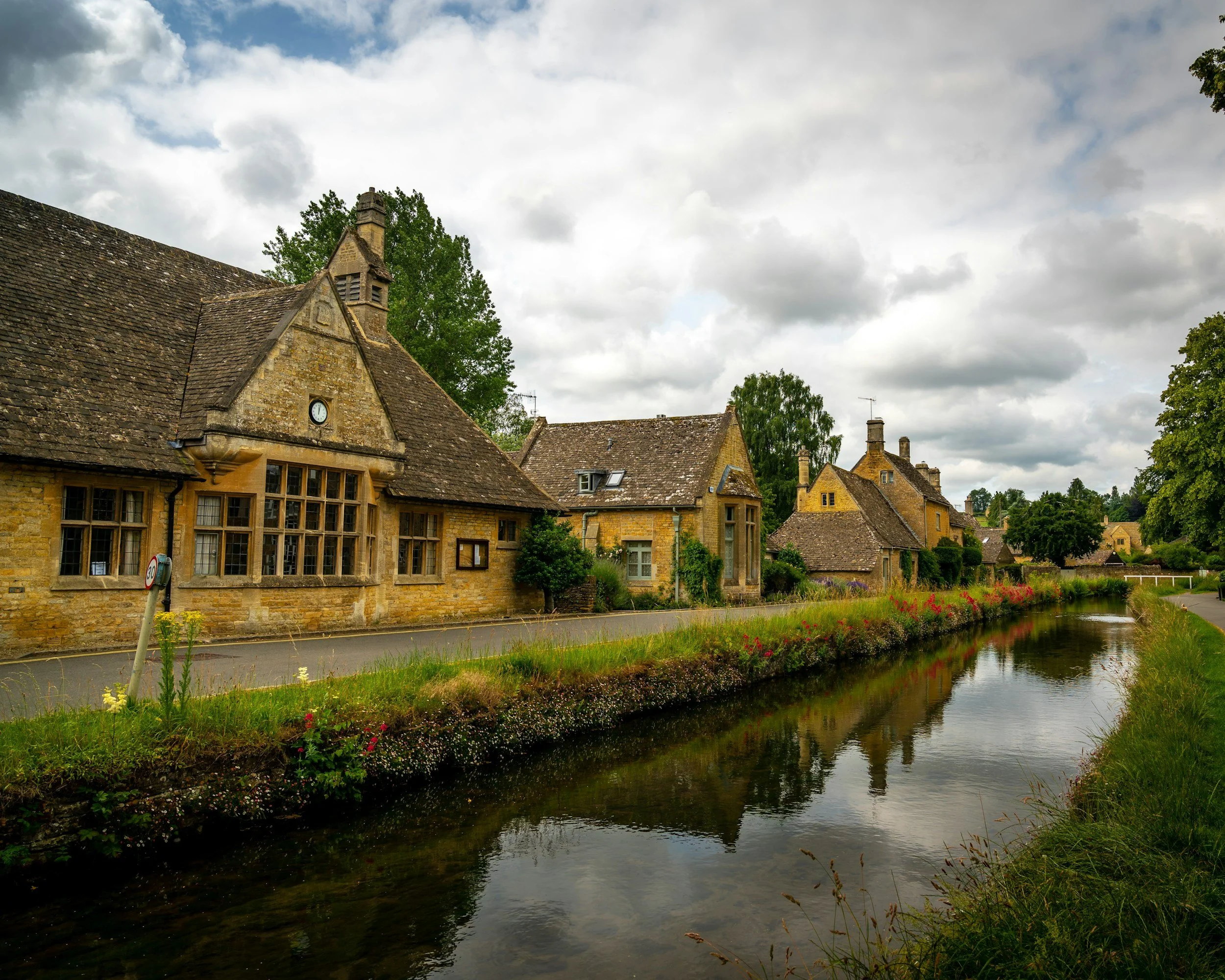 Charming historic village with stone cottages along a small river, lush greenery, and a cloudy sky.