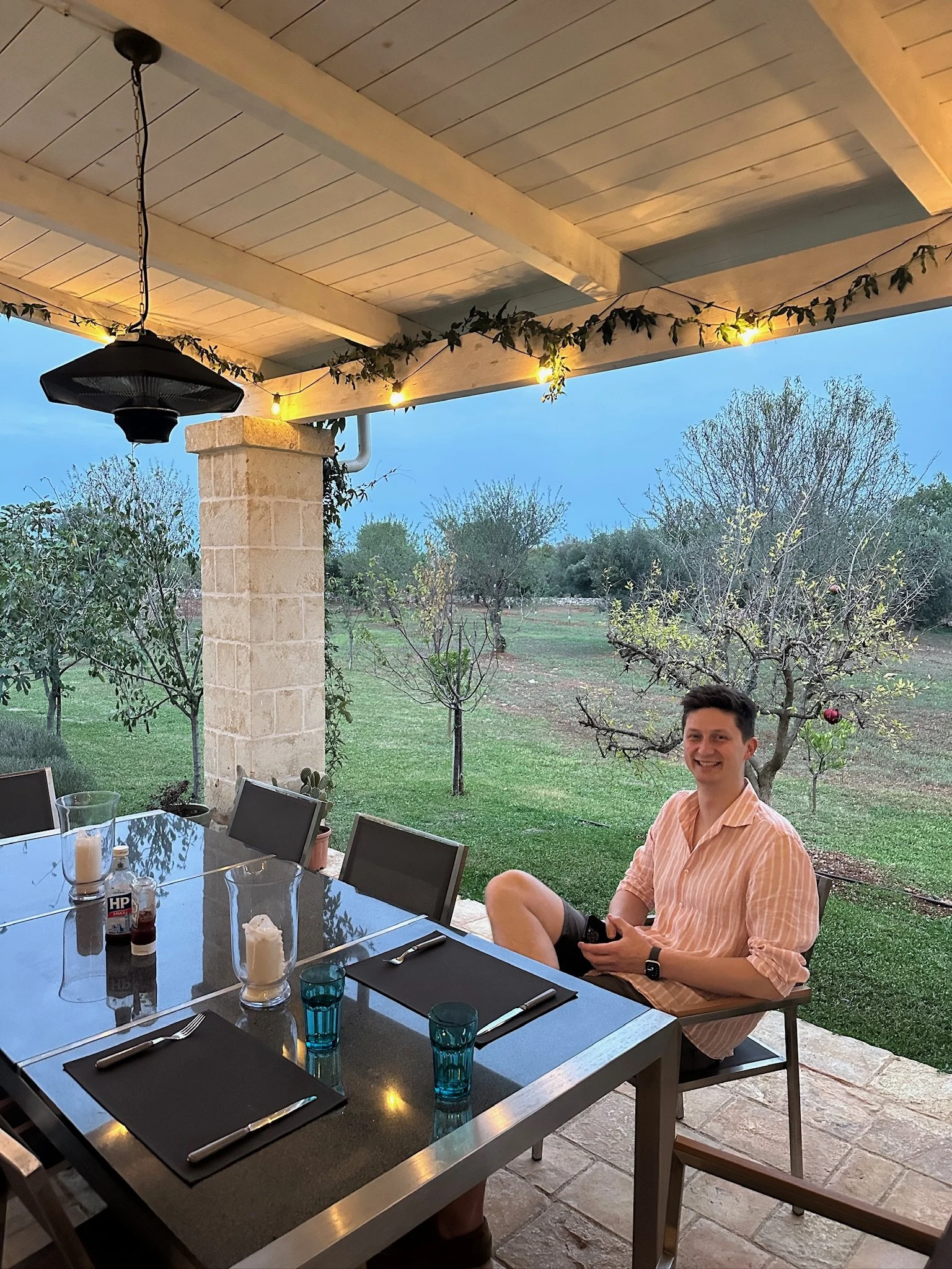A smiling man sitting outdoors at a dining table under a covered patio with string lights, overlooking a backyard with trees and grass.