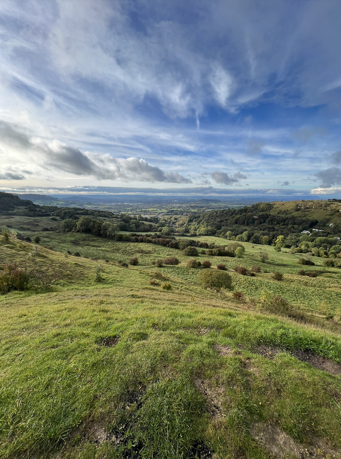 Scenic view of green rolling hills with scattered trees, a valley in the distance, and a partly cloudy sky overhead.