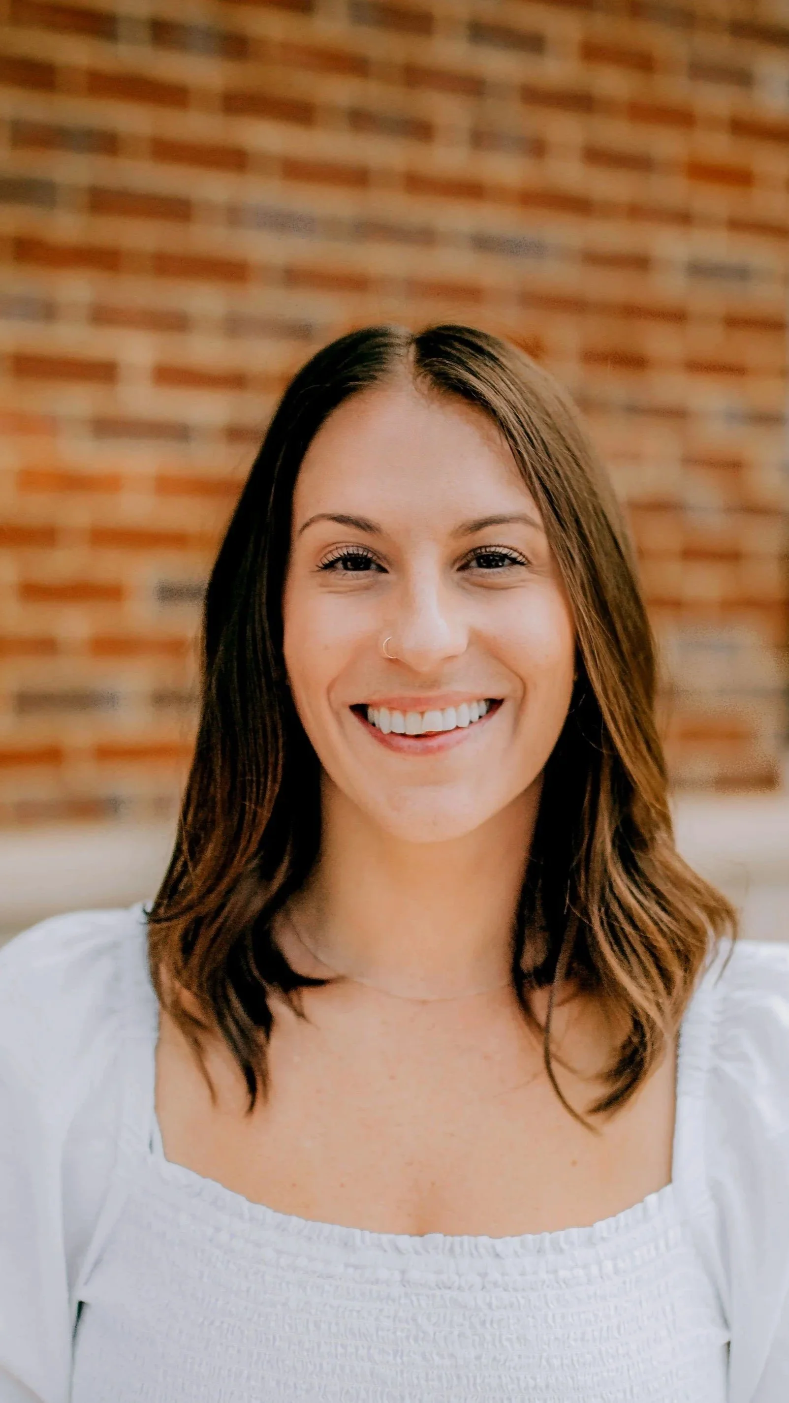 Portrait of a smiling young woman with shoulder-length brown hair, wearing a white top, standing in front of a brick wall.