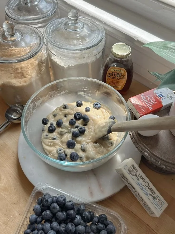 A bowl of blueberry muffin batter being mixed with blueberries. Surrounding items include jars of flour and sugar, honey, milk, and baking ingredients, indicating a baking scene.