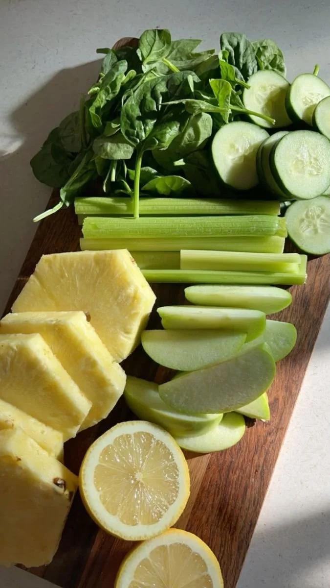 Assorted fresh fruits and vegetables on a wooden cutting board, including pineapple chunks, lemon slices, cucumber slices, celery sticks, apple slices, and leafy greens.