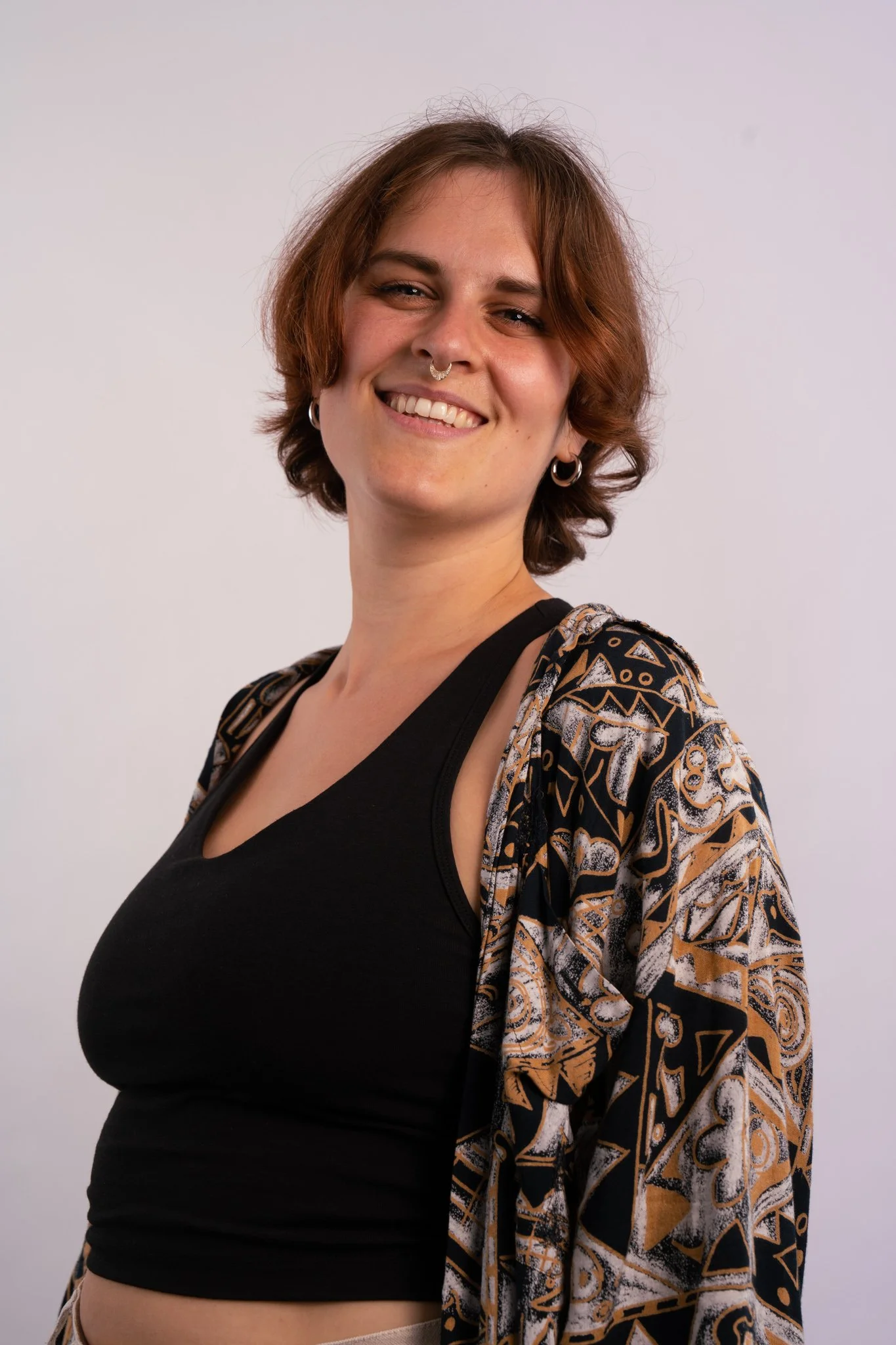 A smiling woman with short brown hair, earrings, and a septum piercing, wearing a black crop top and a patterned jacket, standing against a plain white background.