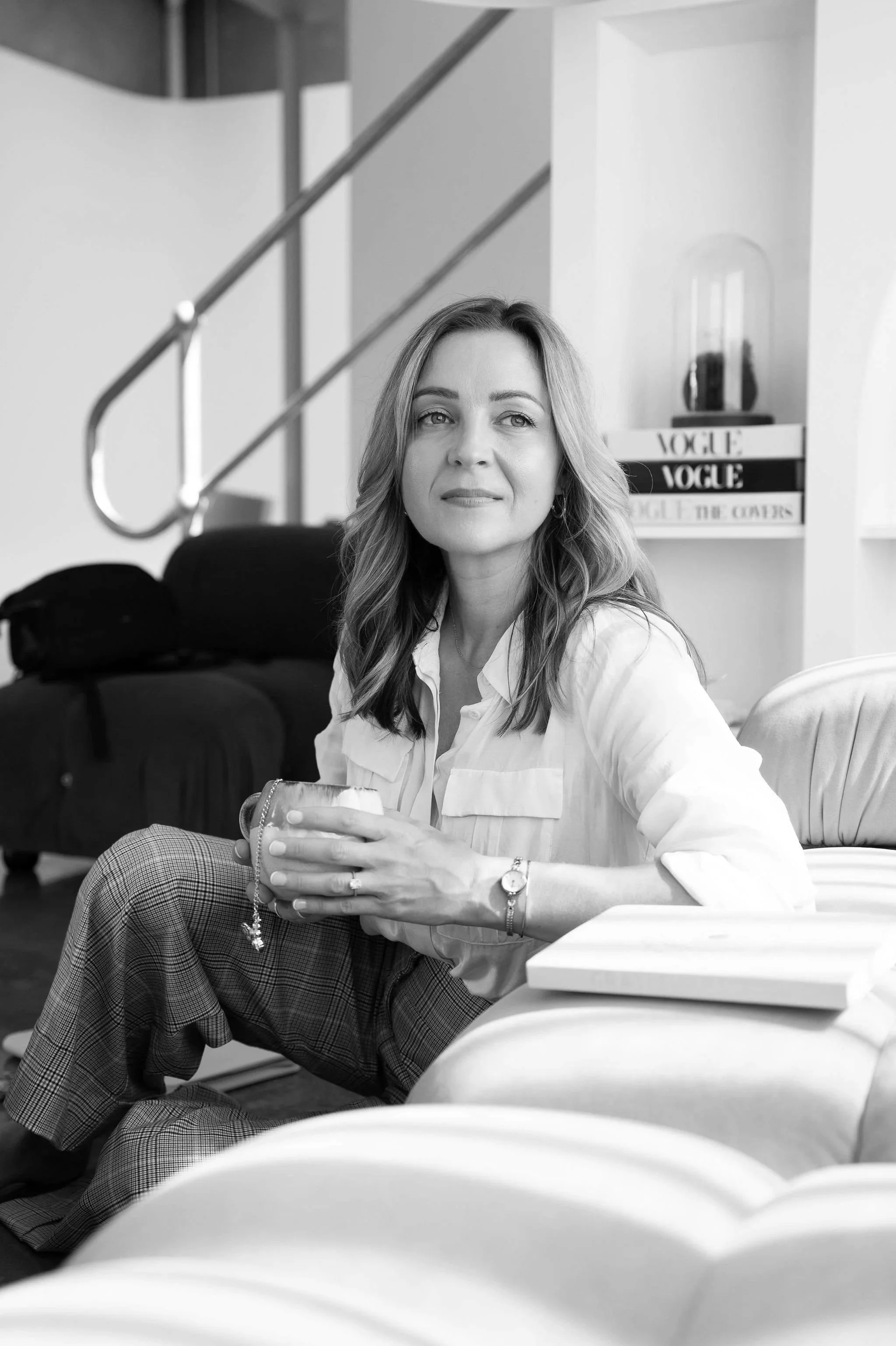 A woman with wavy hair wearing a white blouse and plaid pants sitting on the floor in a modern indoor space, holding a drink in her right hand, with a bookshelf and a decorative object in the background.