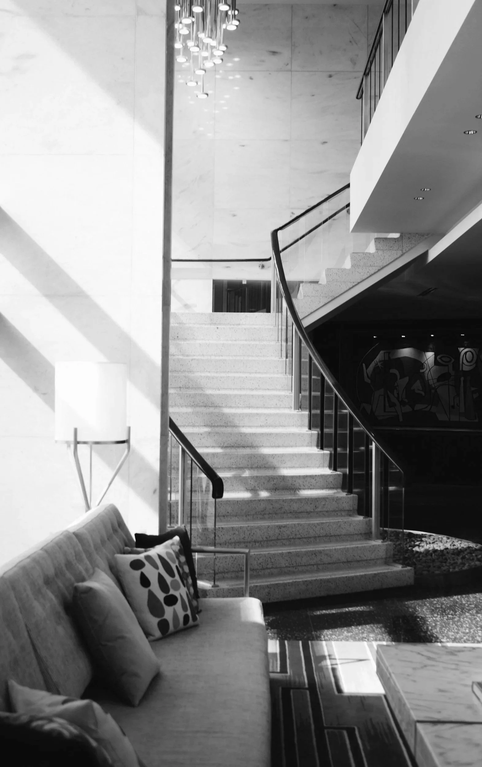 Interior of a modern hotel lobby featuring a staircase with glass railings, a beige sofa with decorative pillows, a side lamp, and contemporary artwork.