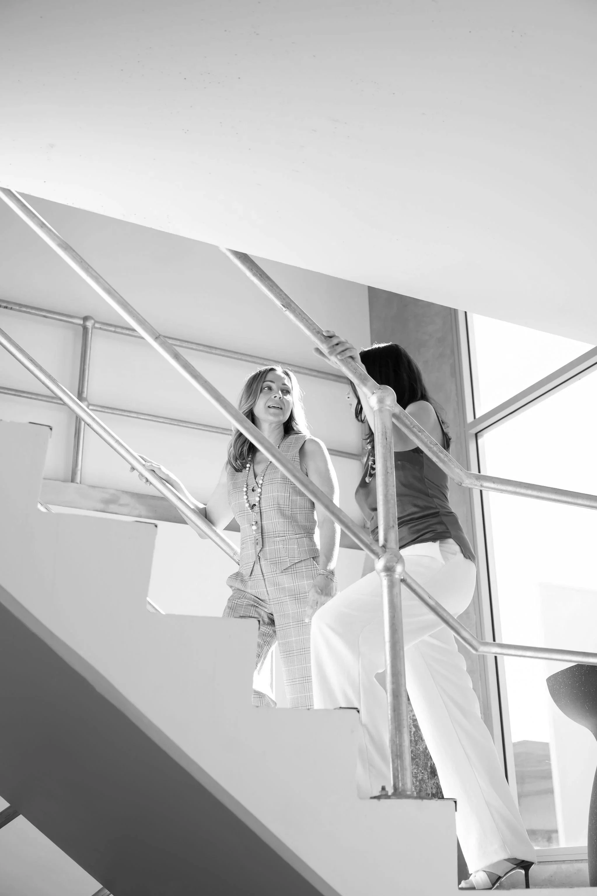 Two women standing on a staircase, engaged in conversation, with one woman wearing a checkered suit and the other wearing a sleeveless top and white pants, illuminated by natural light from a window.
