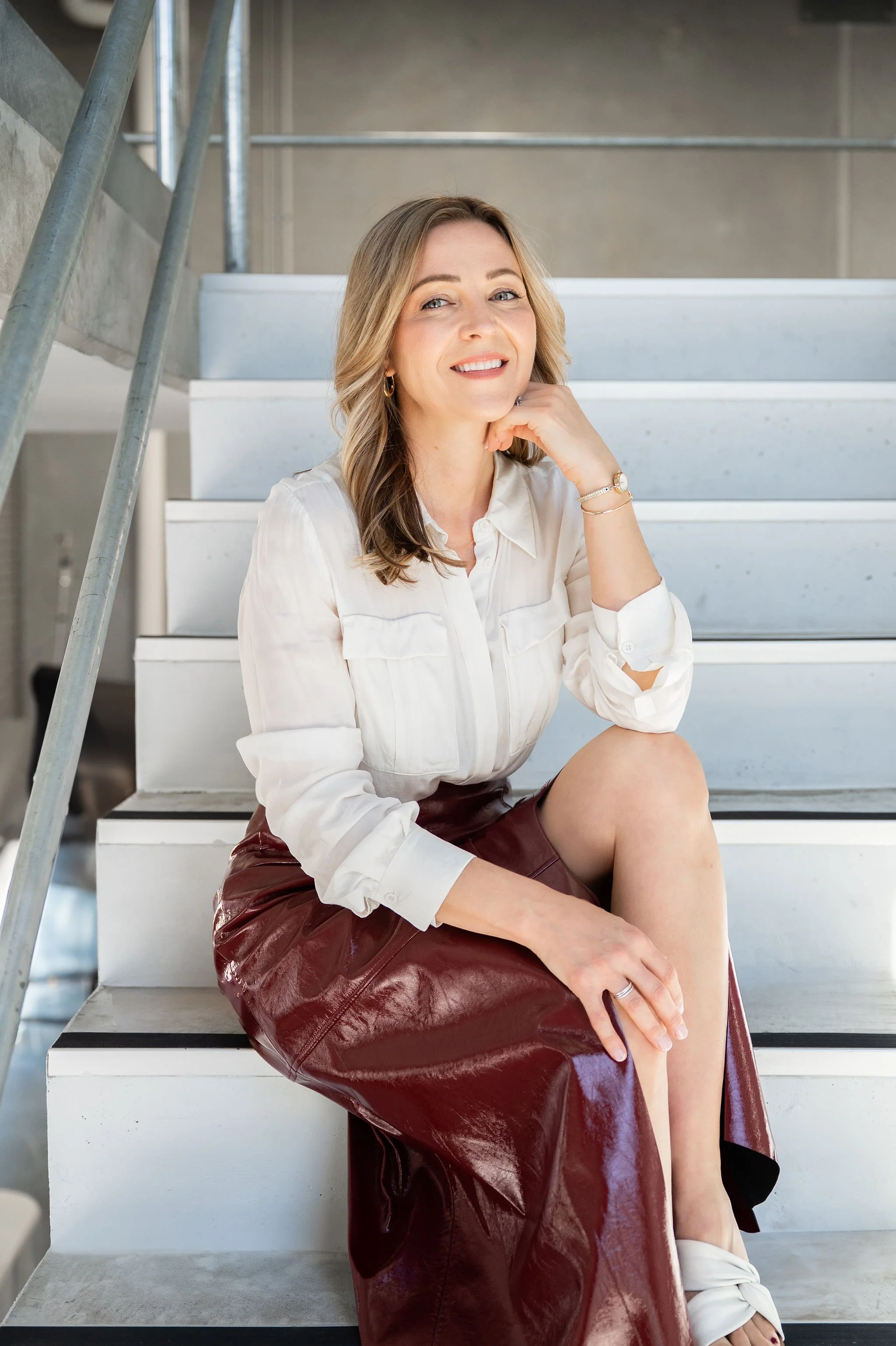 A woman with blonde hair sitting on white stairs in an industrial-style space, wearing a white blouse, burgundy leather skirt, and white shoes, smiling and resting her chin on her hand.