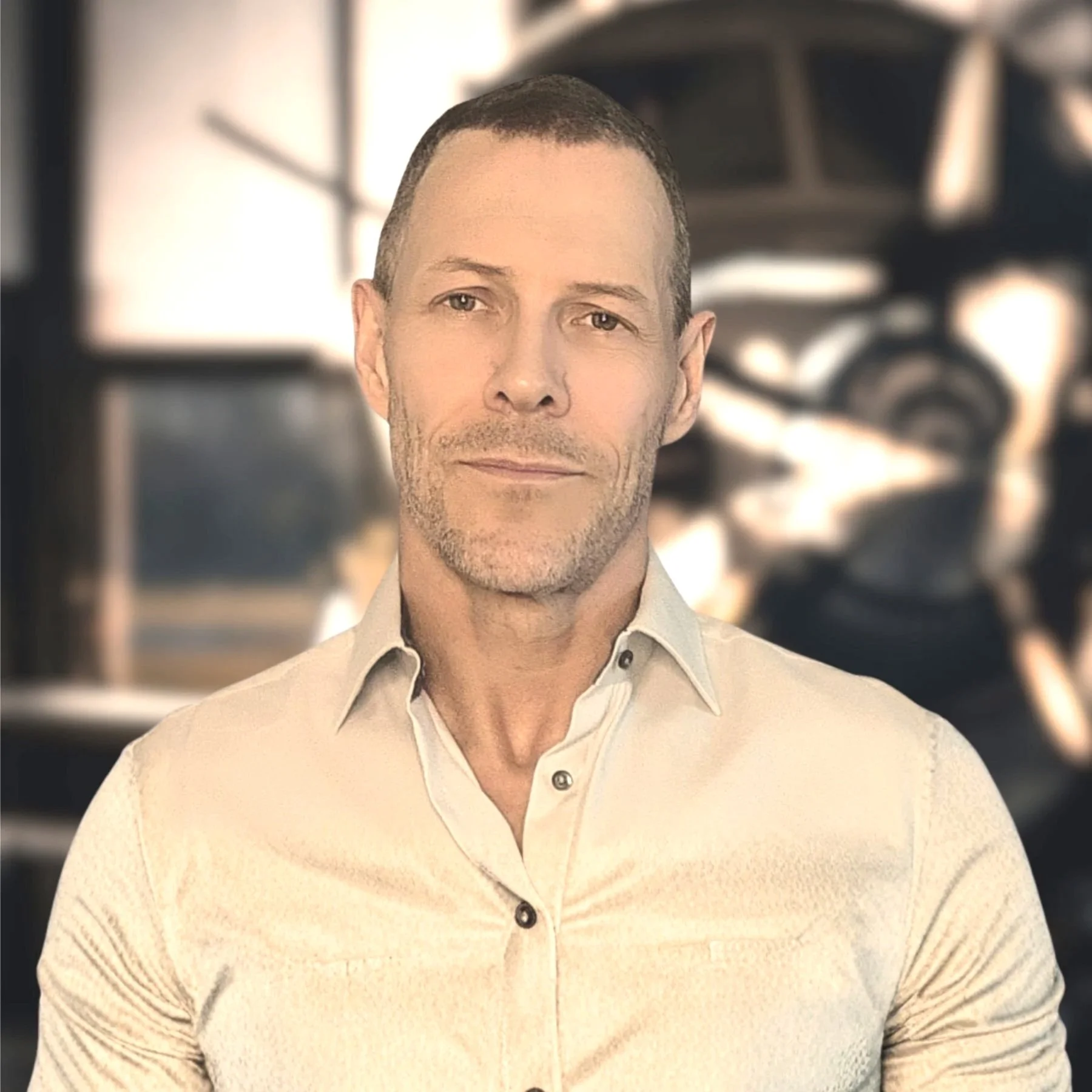 A middle-aged man with short dark hair, lightly stubbled face, wearing a light-colored collared shirt, looking at the camera with a slight smile, with a blurred background of an industrial setting with large machinery and metal beams.