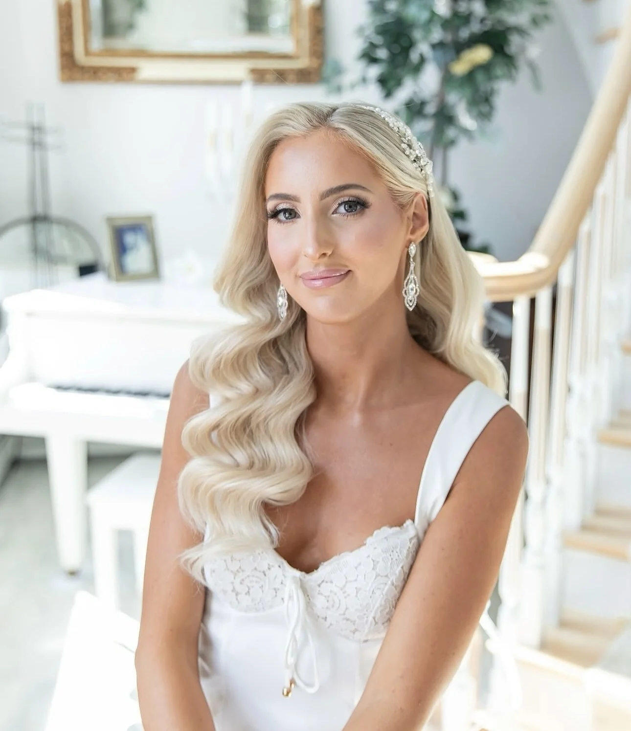 Brunette bride with blonde hair styled in loose waves, wearing a white dress with lace detail, pearl earrings, and a pearl hair accessory, sitting indoors near a staircase with a white piano and framed photographs in the background.