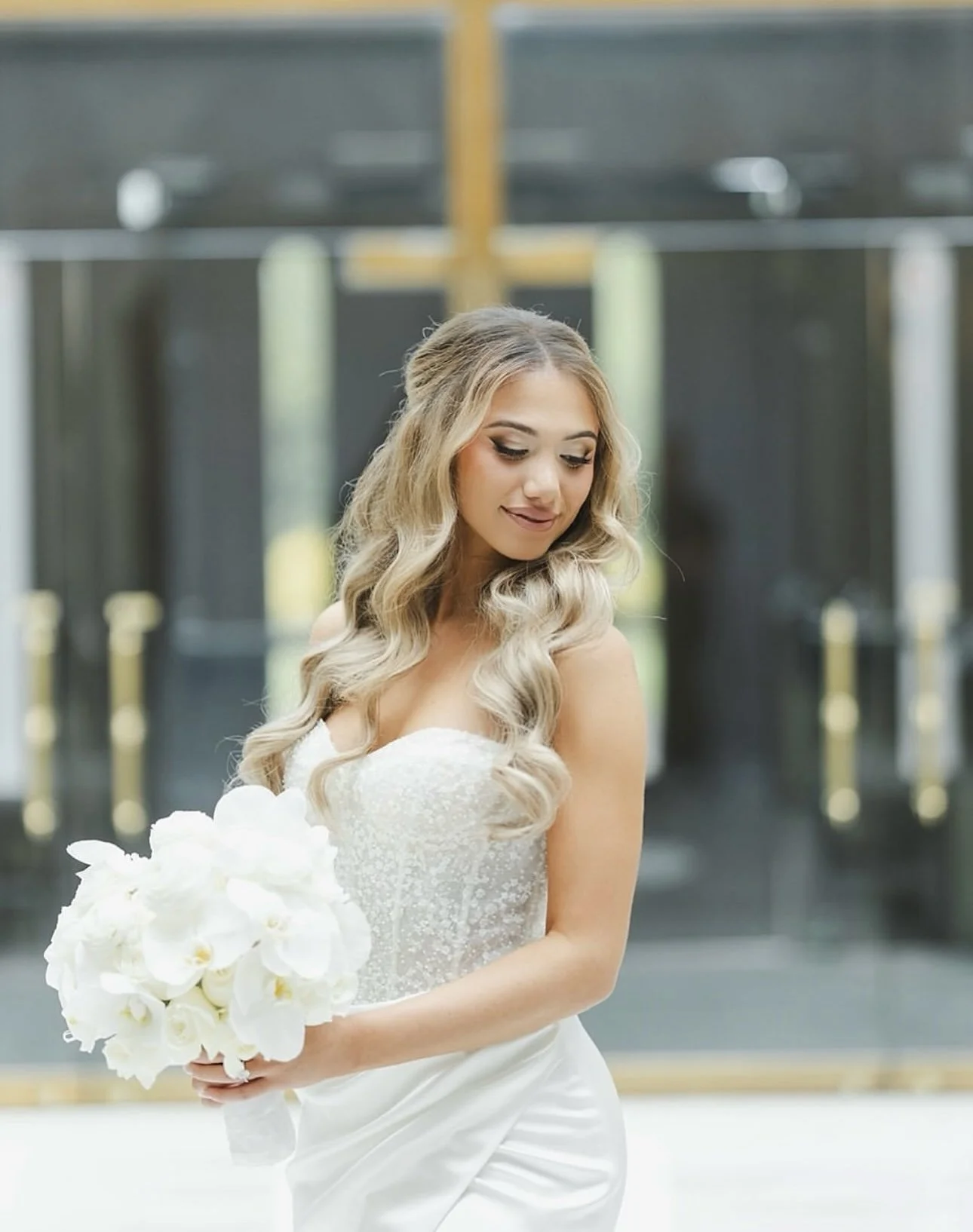 A bride in a wedding dress holding a bouquet of white flowers, standing in front of glass doors.