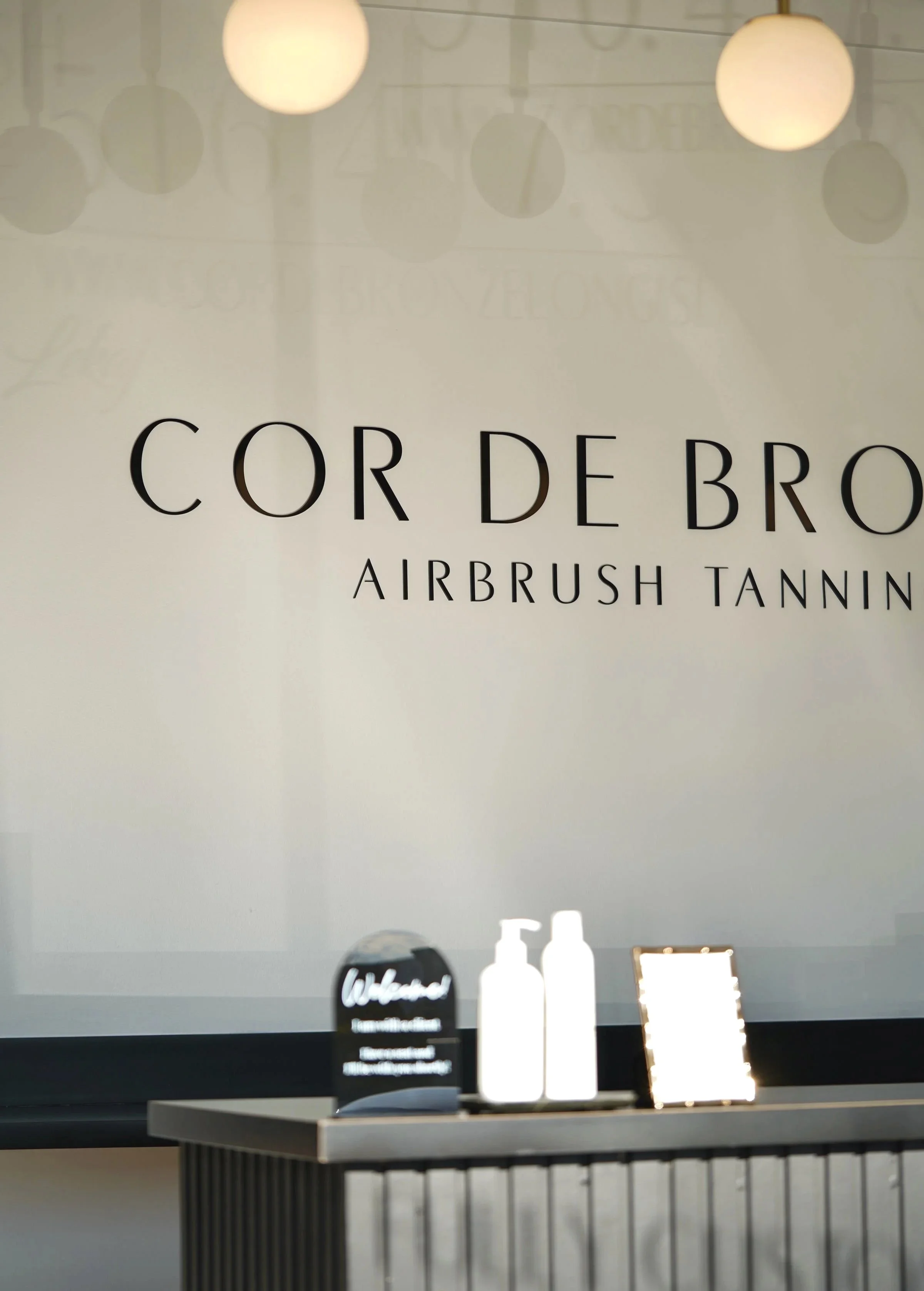 Display shelf with bottles and a small sign, logo in the background for a salon or beauty business, with round lights hanging from the ceiling.