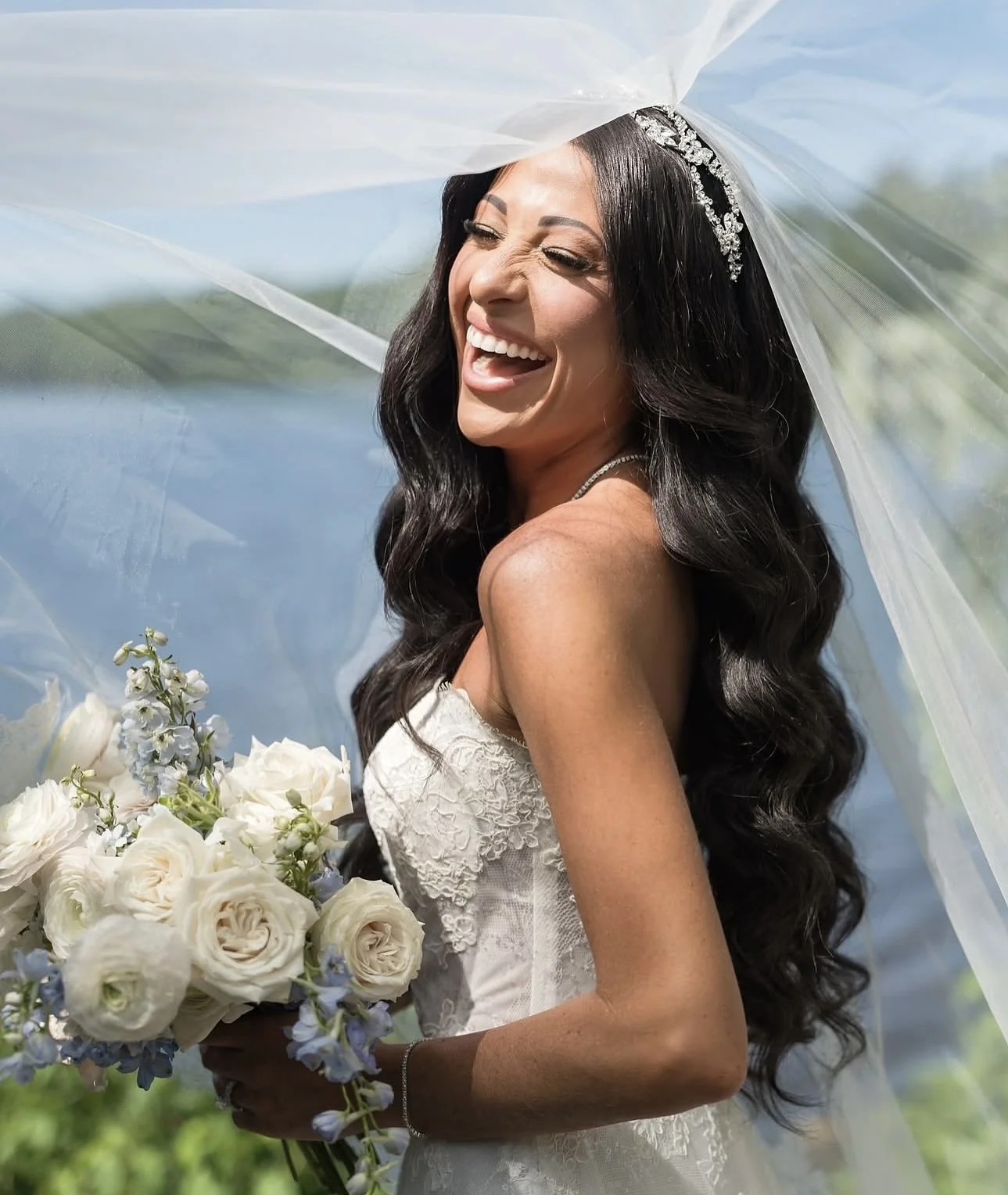 A smiling bride with long dark hair and a veil, holding a bouquet of white roses and other flowers.
