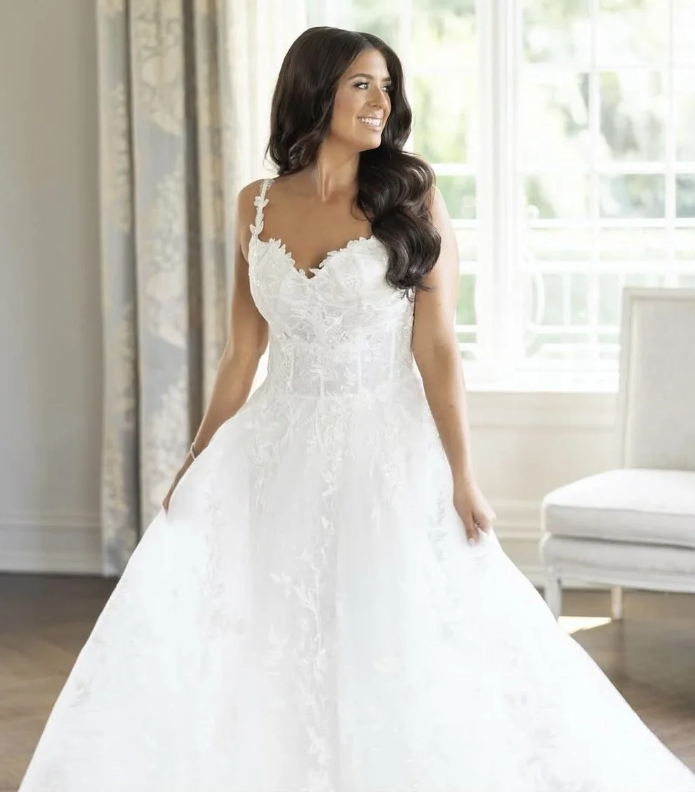 A woman in a white wedding dress standing indoors near a window with patterned curtains, smiling and looking to the side.