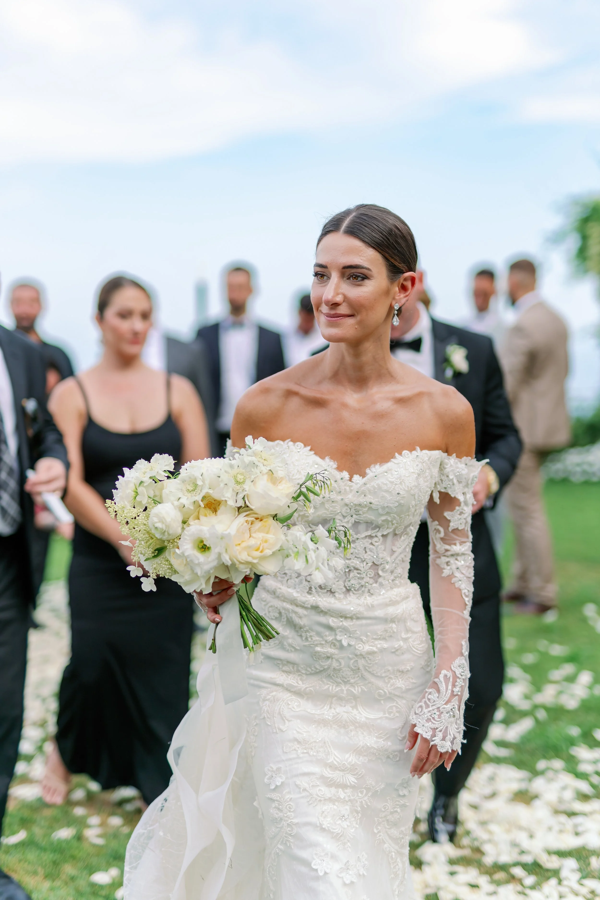 Bride in an off-shoulder white wedding dress holding a bouquet of white flowers at an outdoor wedding reception.