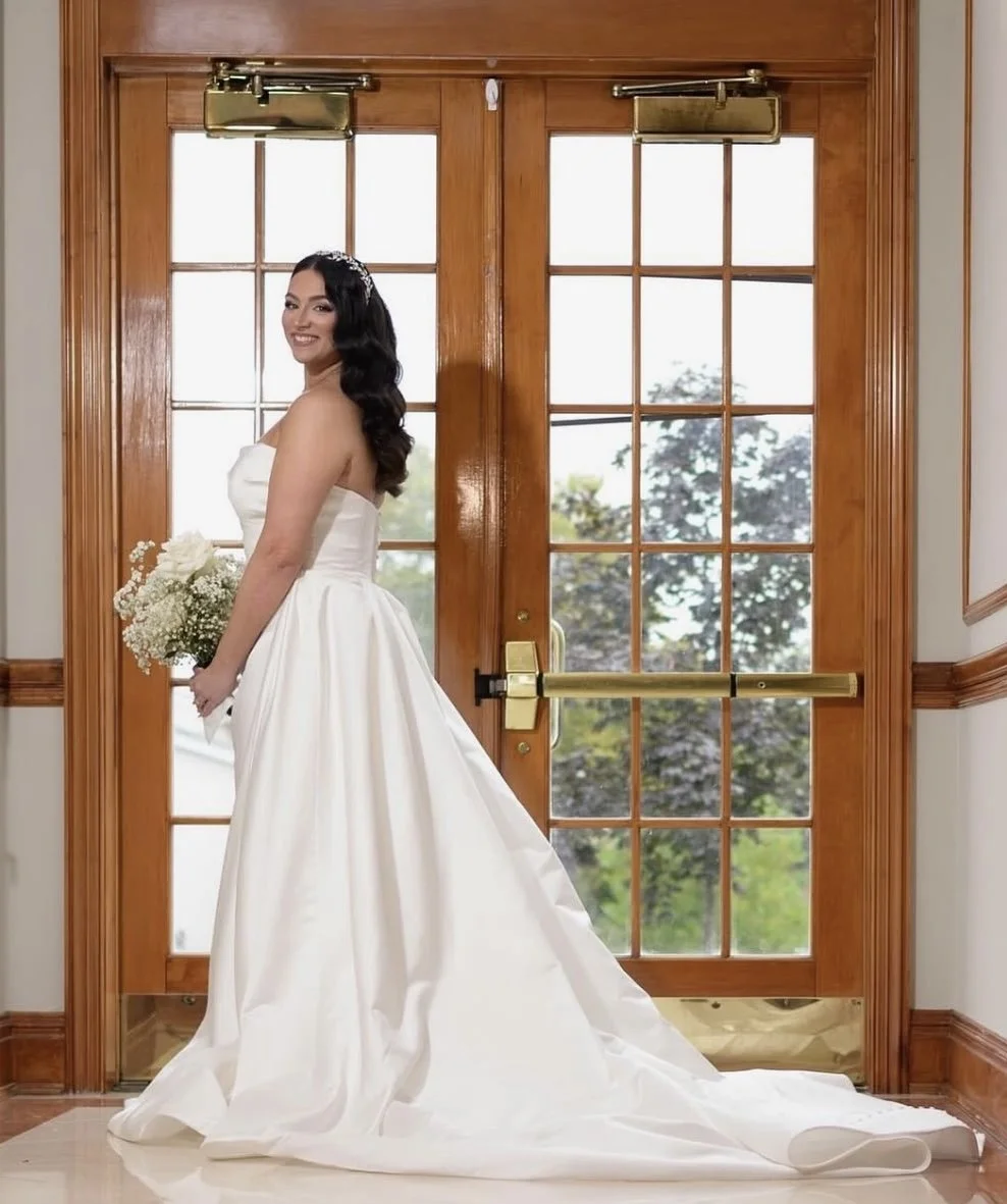 A bride in a white wedding gown standing in front of wooden double doors with glass panels, holding a bouquet of white flowers, smiling.
