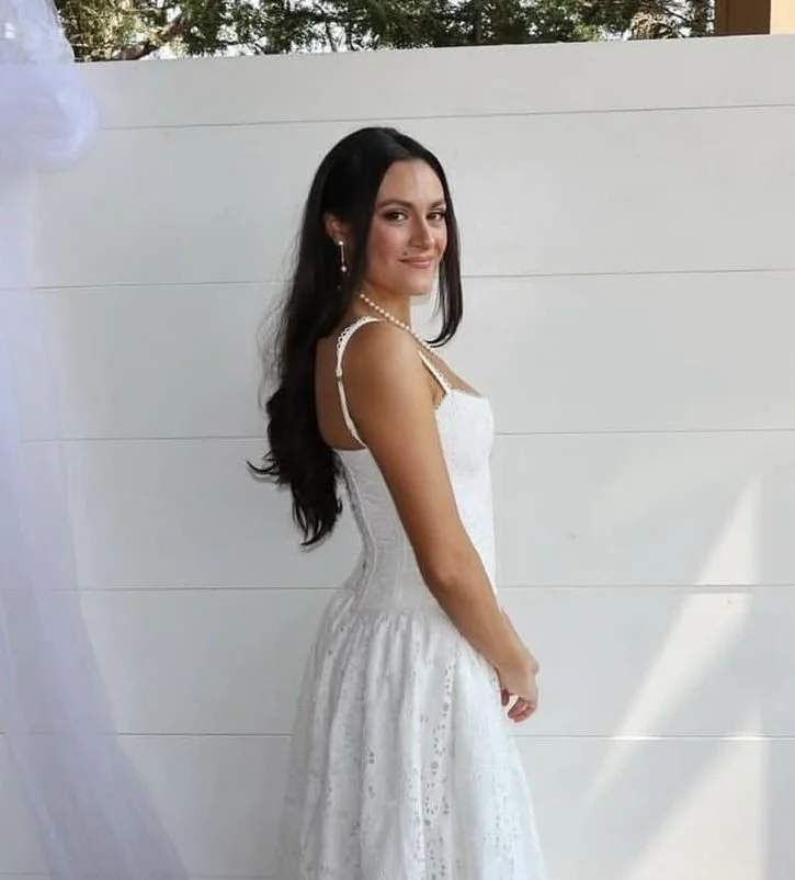 A woman with long dark hair in a white dress standing outdoors against a white wall.