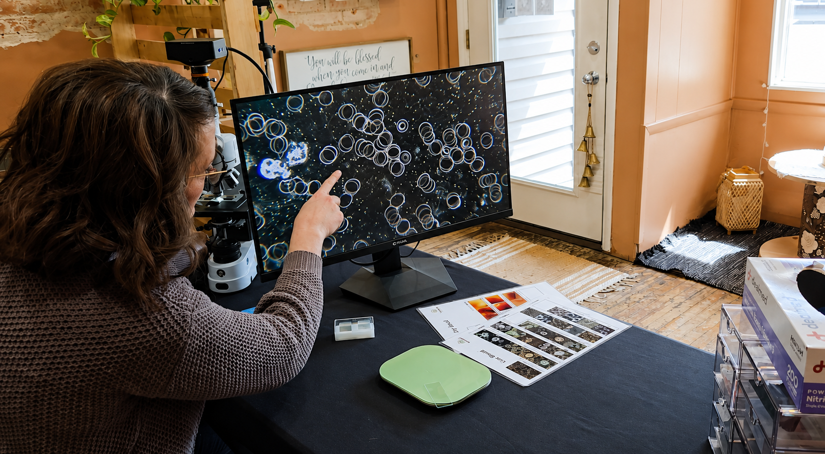 A woman with curly hair wearing glasses and a brown sweater is pointing at a computer monitor displaying a microscopic image of cells or bacteria. The workspace is set up on a black table with scientific materials, including charts, a microscope, and sample slides.