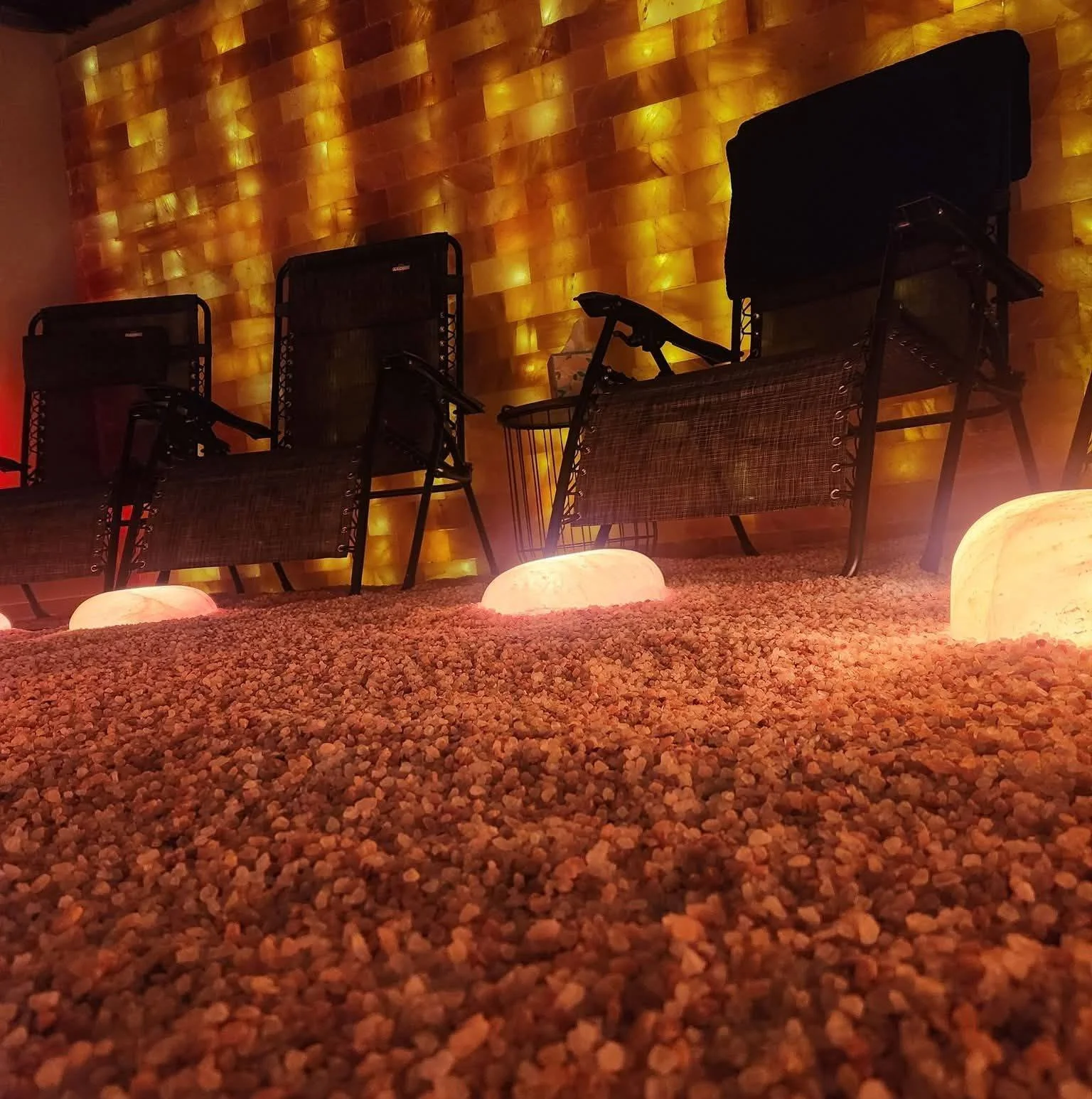 Indoor seating area with three black chairs, illuminated salt lamps, a textured wall with warm backlighting, and a gravel floor.