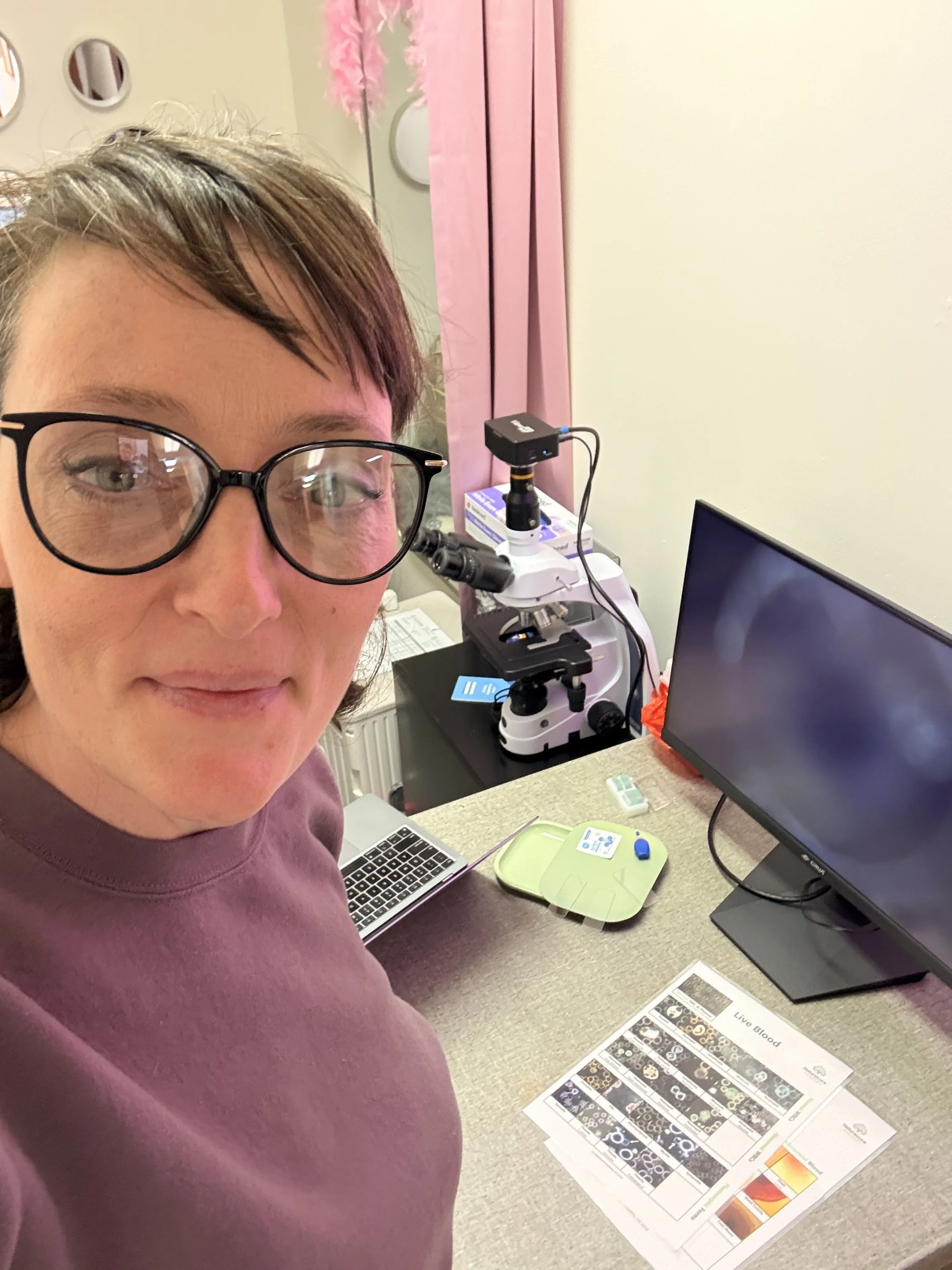 A woman with glasses taking a selfie in a medical or laboratory setting. She is standing next to a microscope, a computer monitor, and various medical supplies on the desk. There is a blood smear chart on the desk.