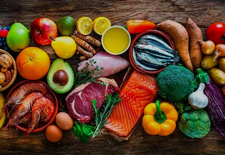 A variety of fresh fruits, vegetables, seafood, and meats arranged on a wooden surface, including apples, oranges, avocado, lemon, shrimp, salmon, steak, broccoli, garlic, sweet potatoes, and eggs.