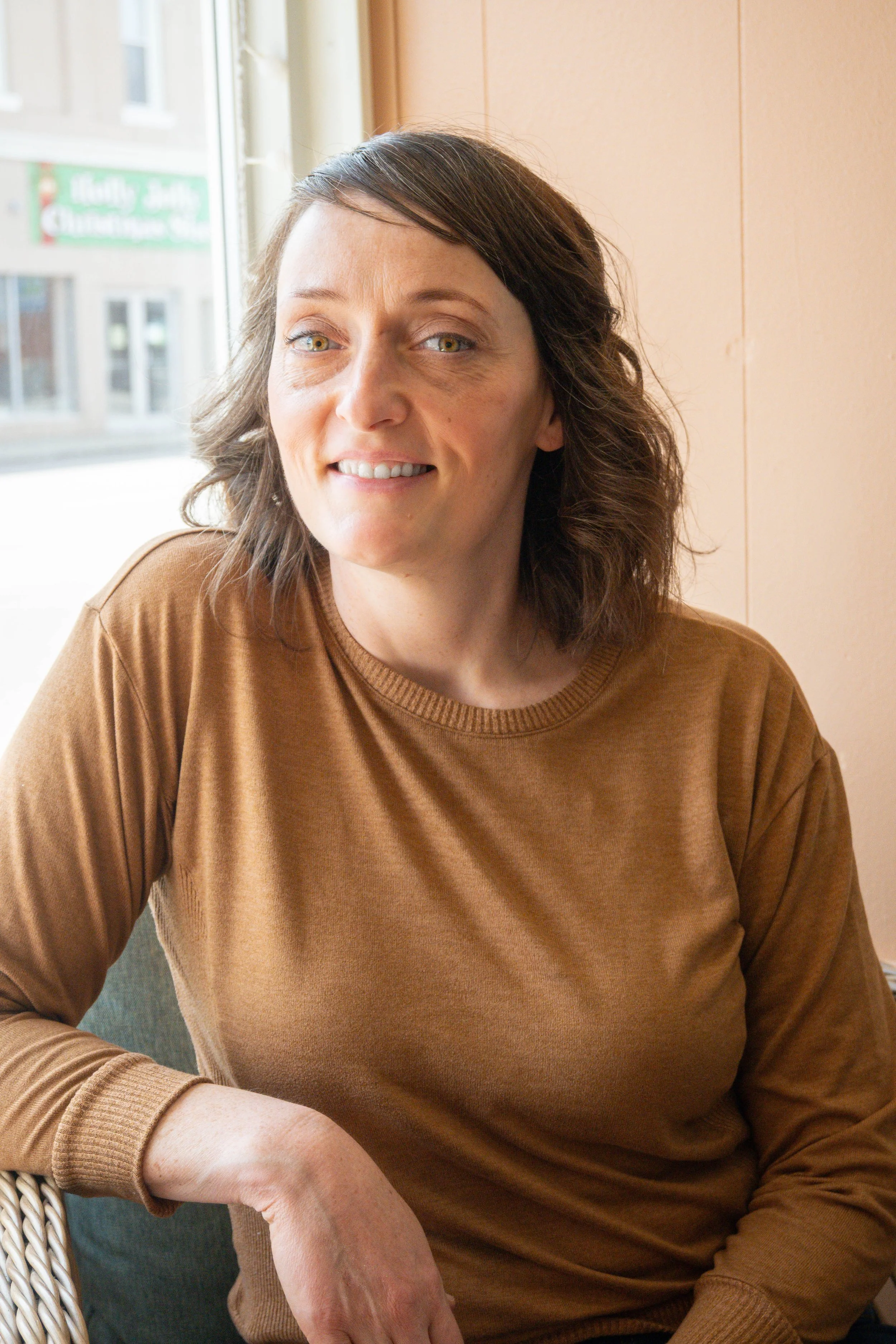 A woman with shoulder-length brown hair and green eyes, wearing a brown long-sleeve shirt, smiling and sitting near a window with city buildings visible outside.